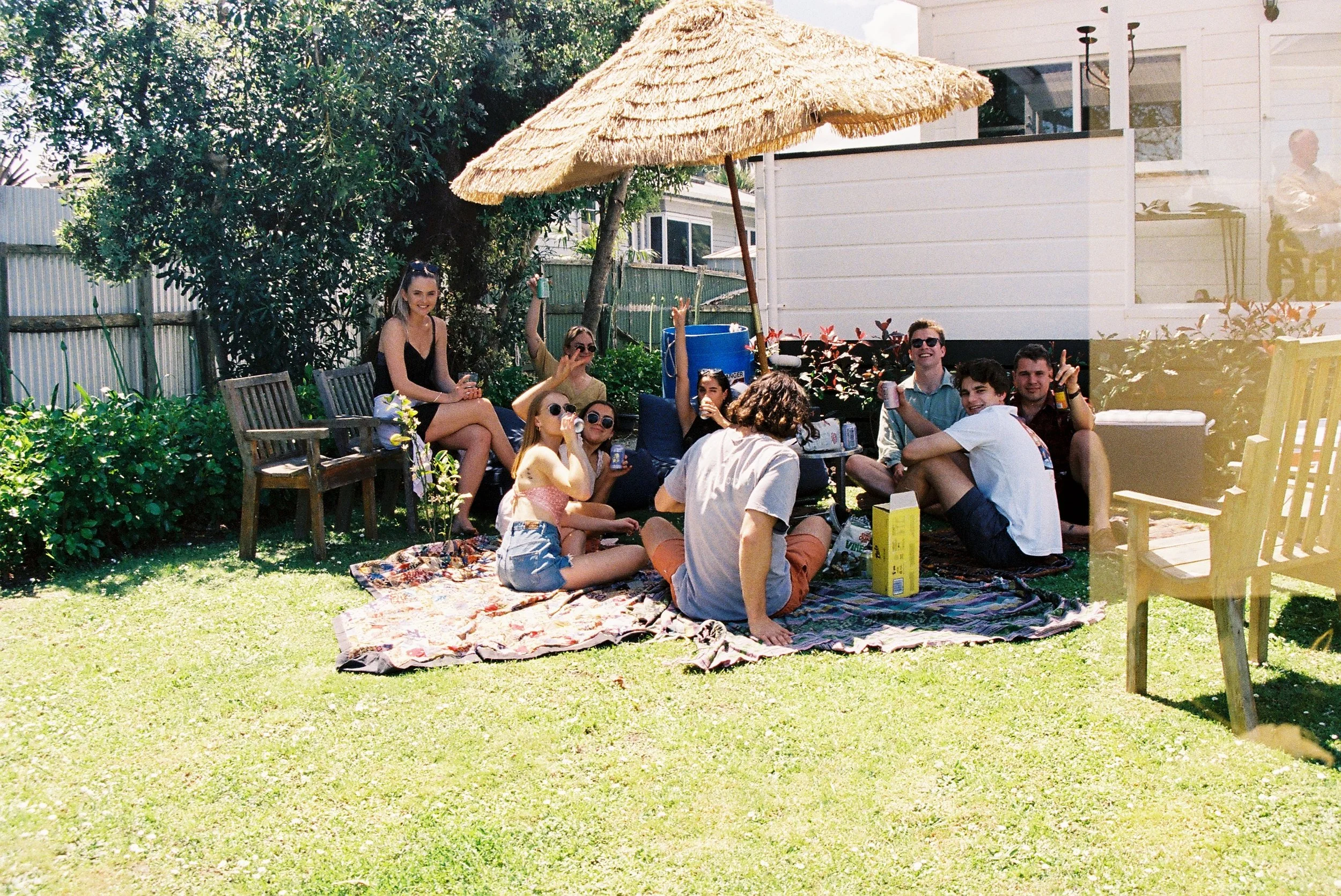 Group of friends gathering outdoors on a sunny day, sitting on a blanket and chairs, under a thatched umbrella in a backyard with green grass and bushes.