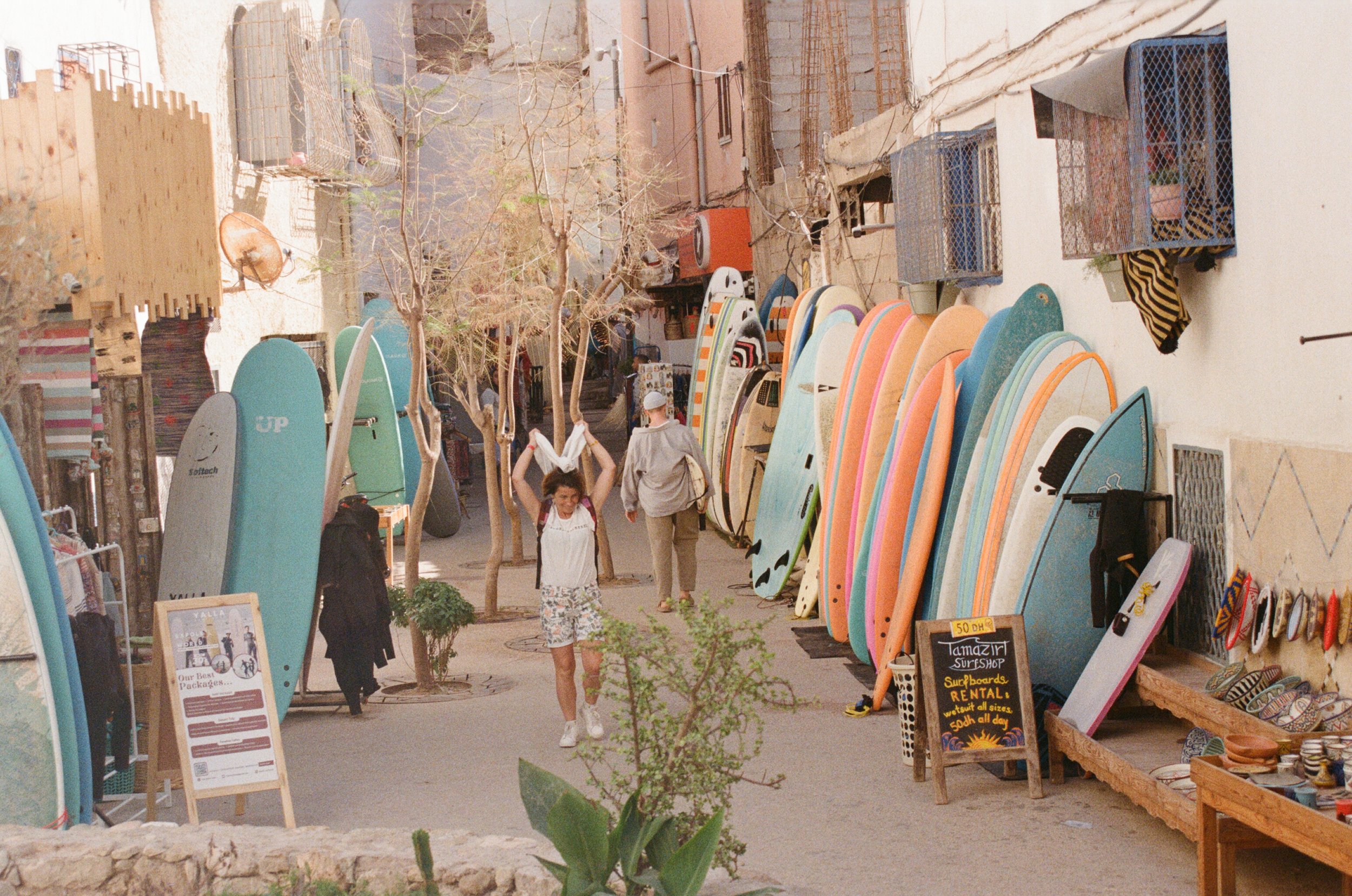 A street lined with colorful surfboards leaning against walls and trees, with a woman wearing a white tank top and shorts walking and smiling, and another person in the background. There are signs and various accessories displayed for sale.