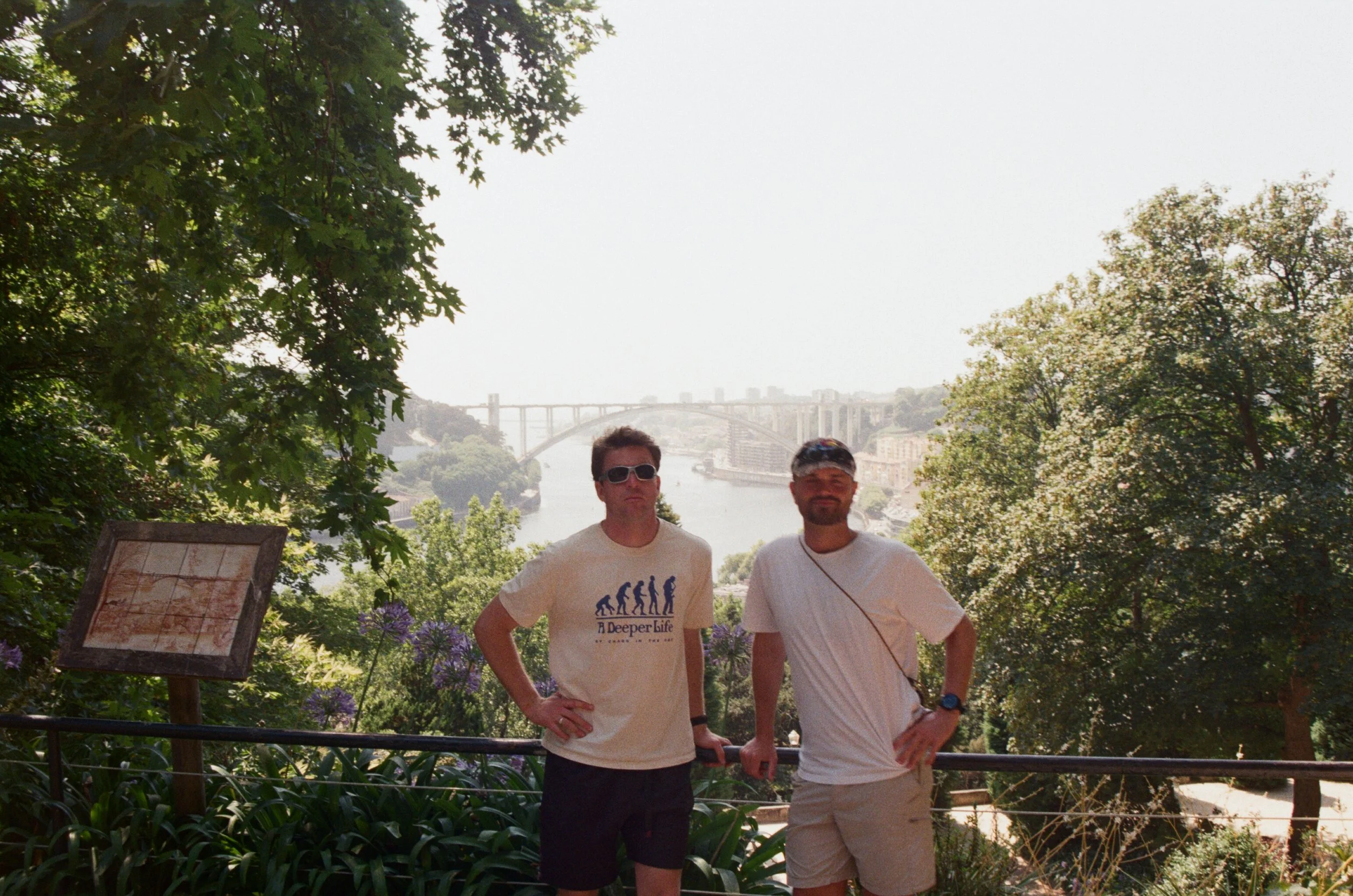 Two men standing outdoors with a city skyline and bridge in the background; one wearing sunglasses and a graphic T-shirt, the other with a beard, cap, and white T-shirt; greenery surrounds them.
