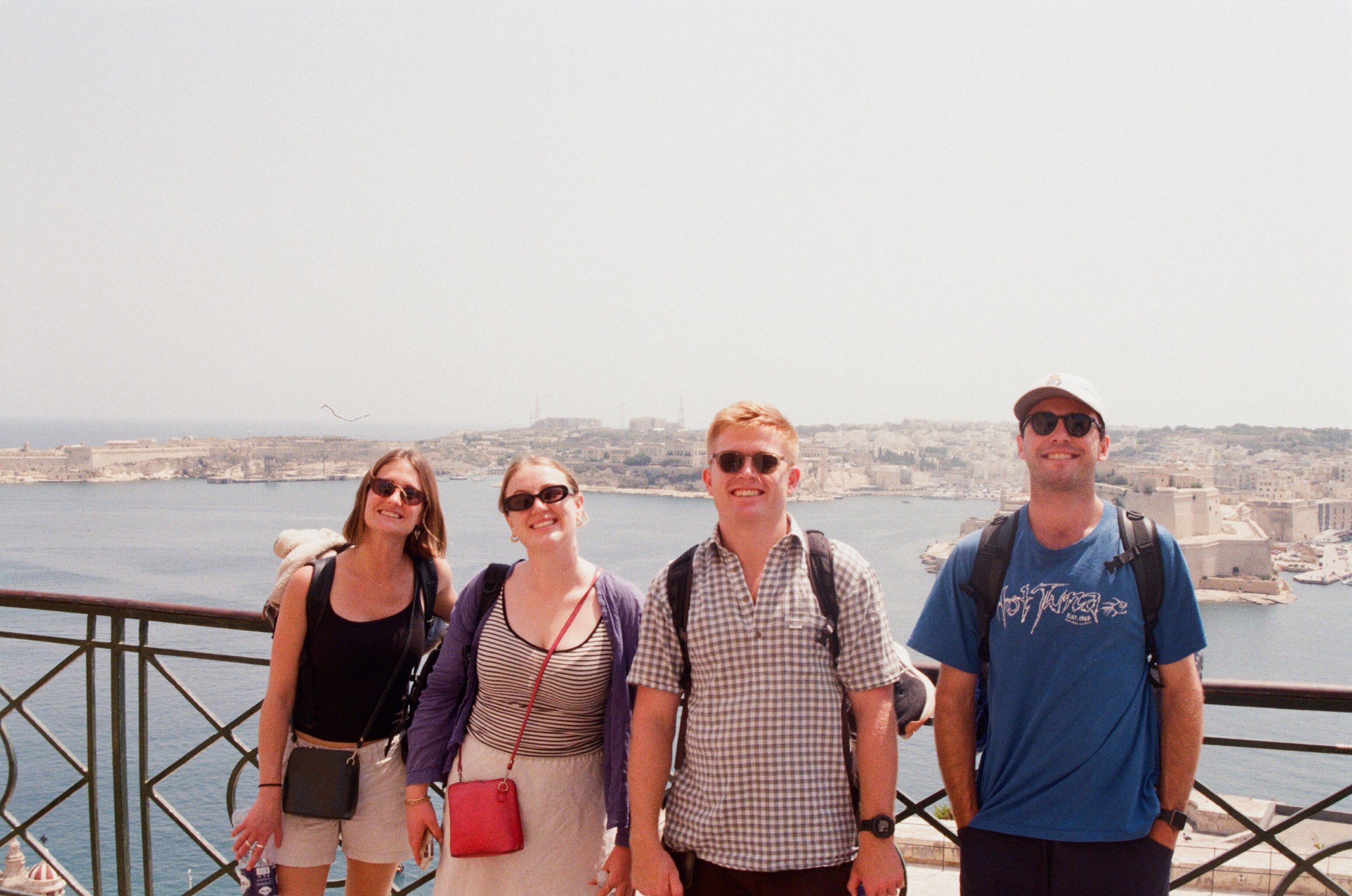 Four friends smiling at the camera on a balcony with a waterfront city and fort in the background.