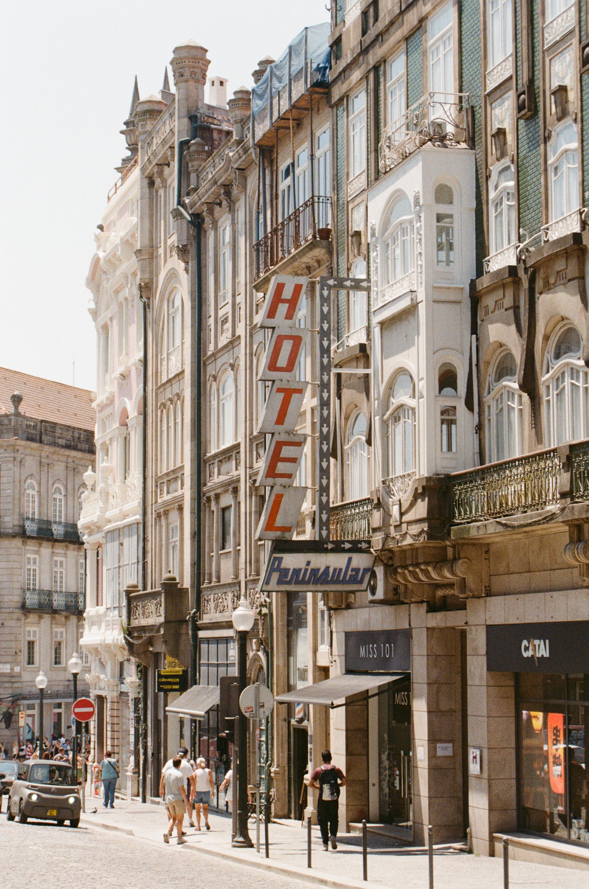 Street view in a European city with historic buildings, pedestrians, cars, and a hotel sign.