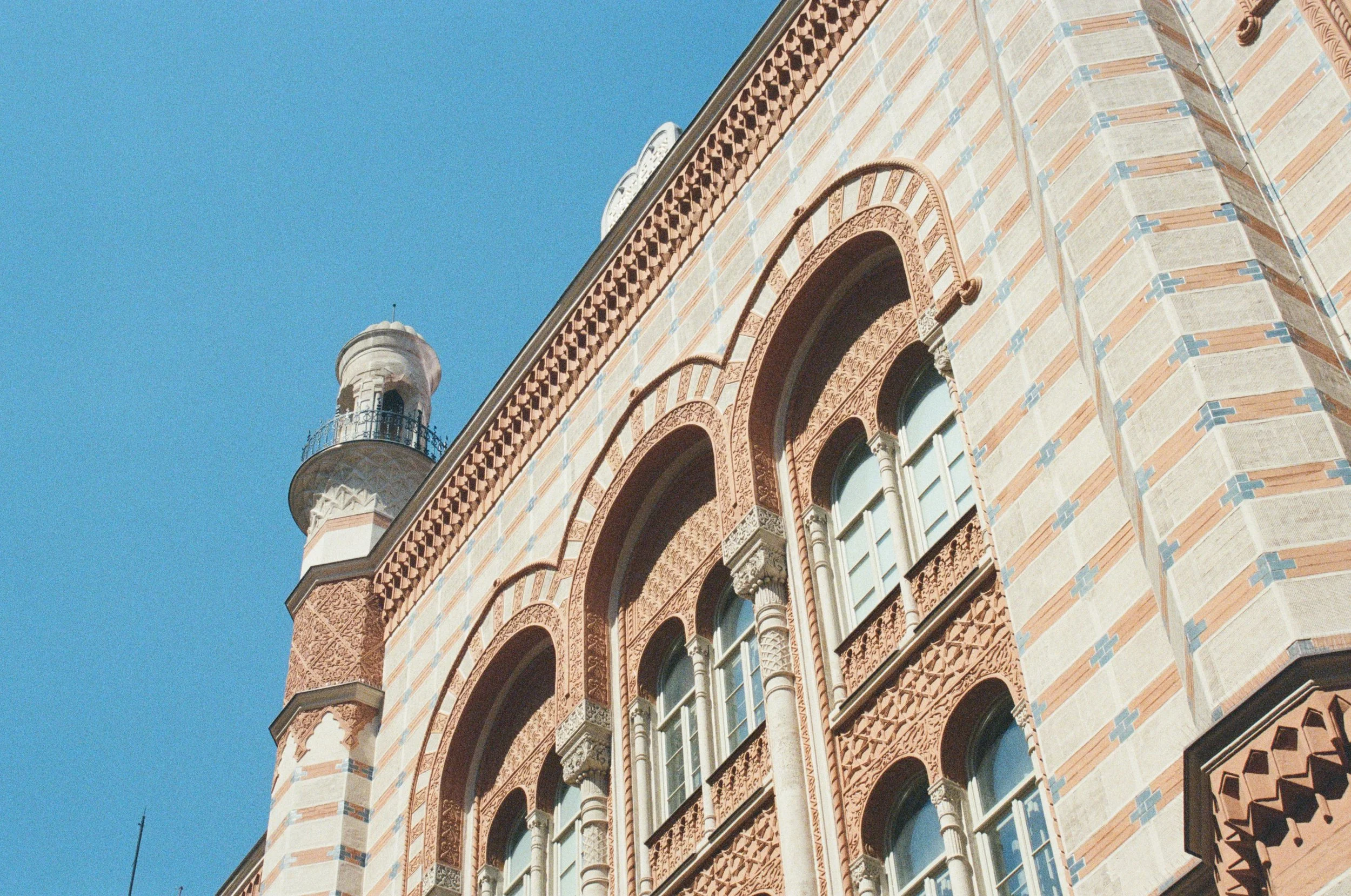 Close-up of a historic building with ornate brickwork, arched windows, and a corner tower with a balcony and a small dome, set against a clear blue sky.