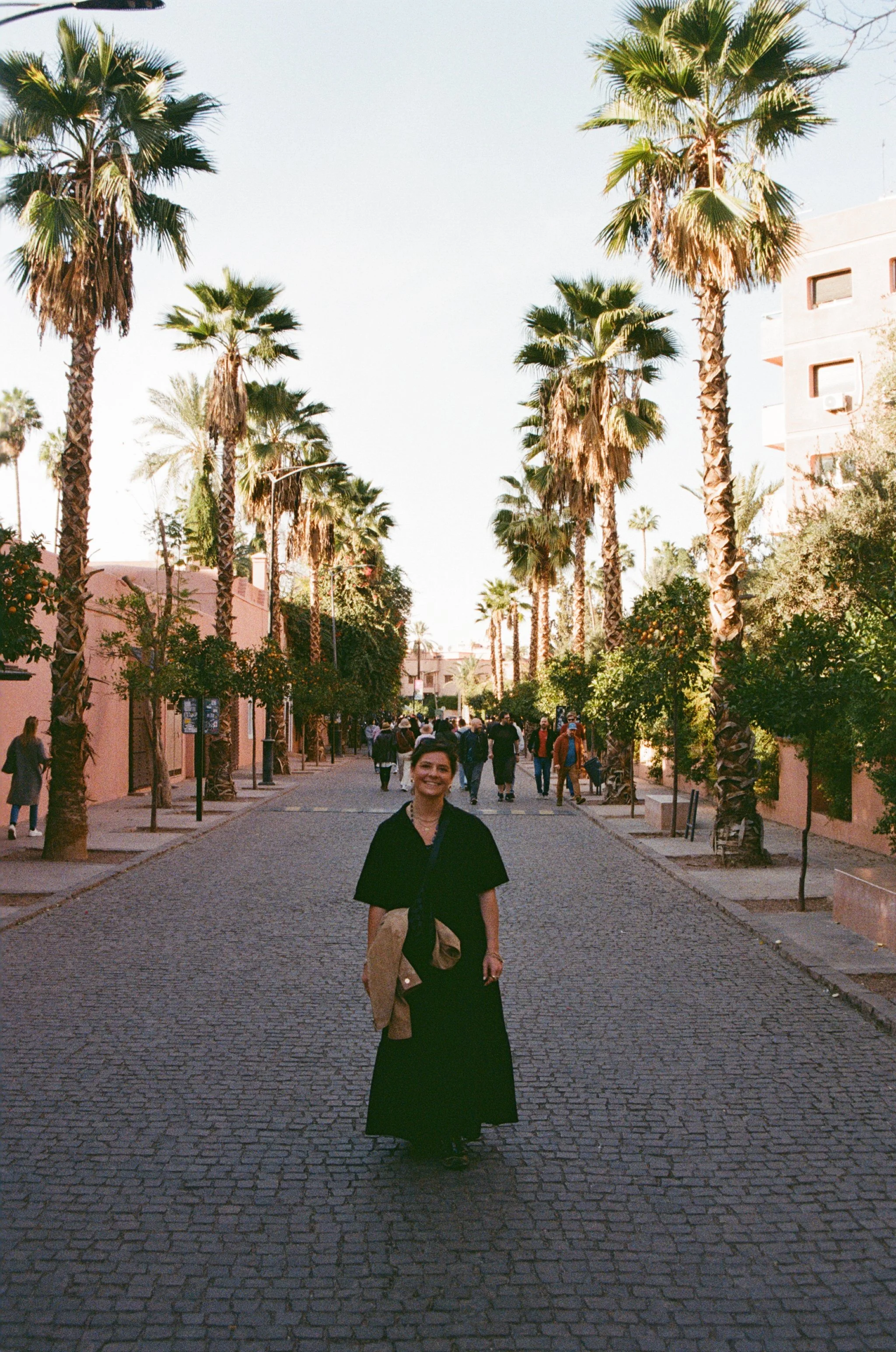 A woman standing on a cobblestone street lined with tall palm trees and pink buildings, smiling at the camera.