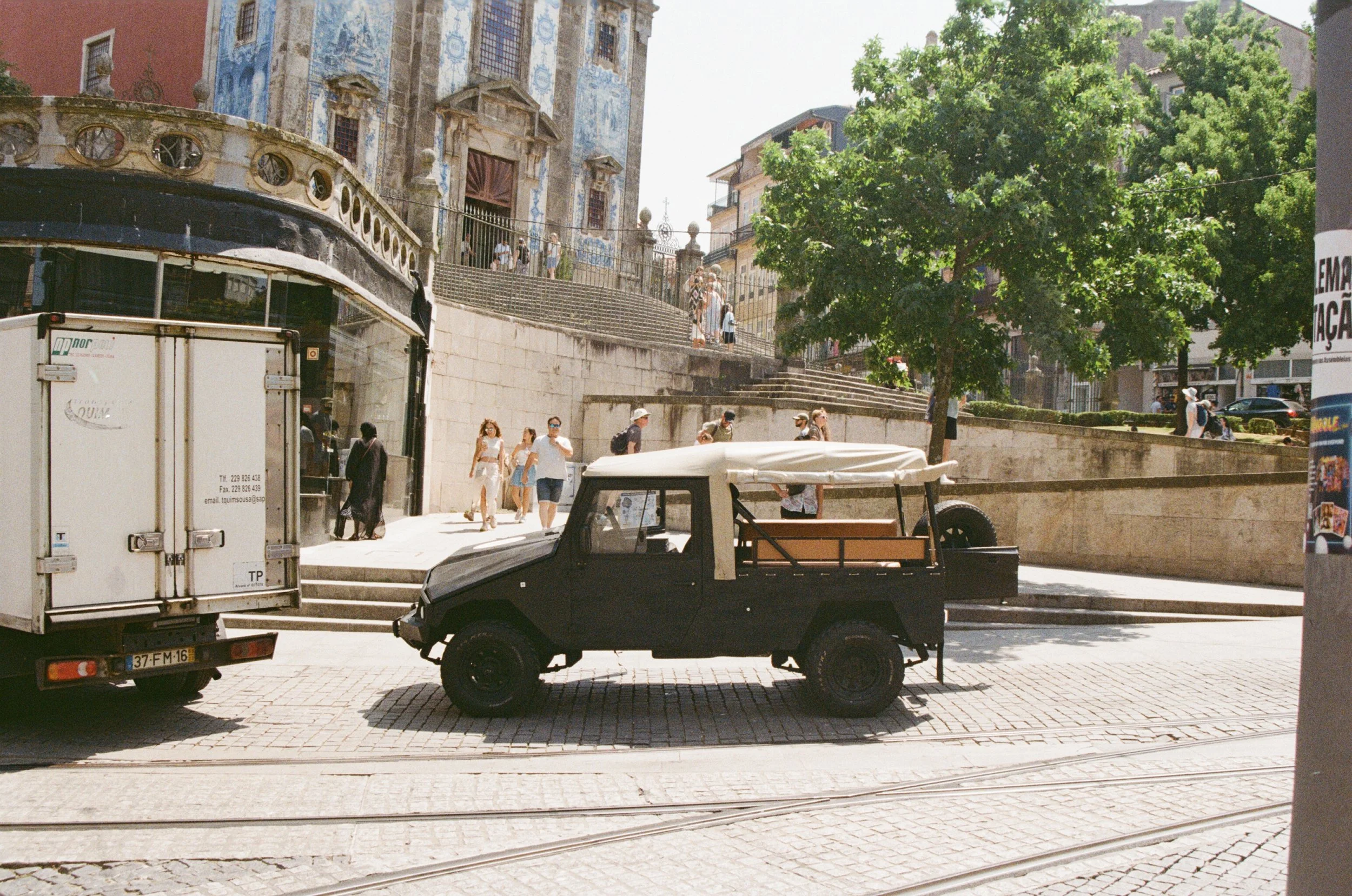 A black utility vehicle parked on a city street near a white delivery truck, with steps leading up to a historic building with blue and gold details in the background, and people walking along the sidewalk.