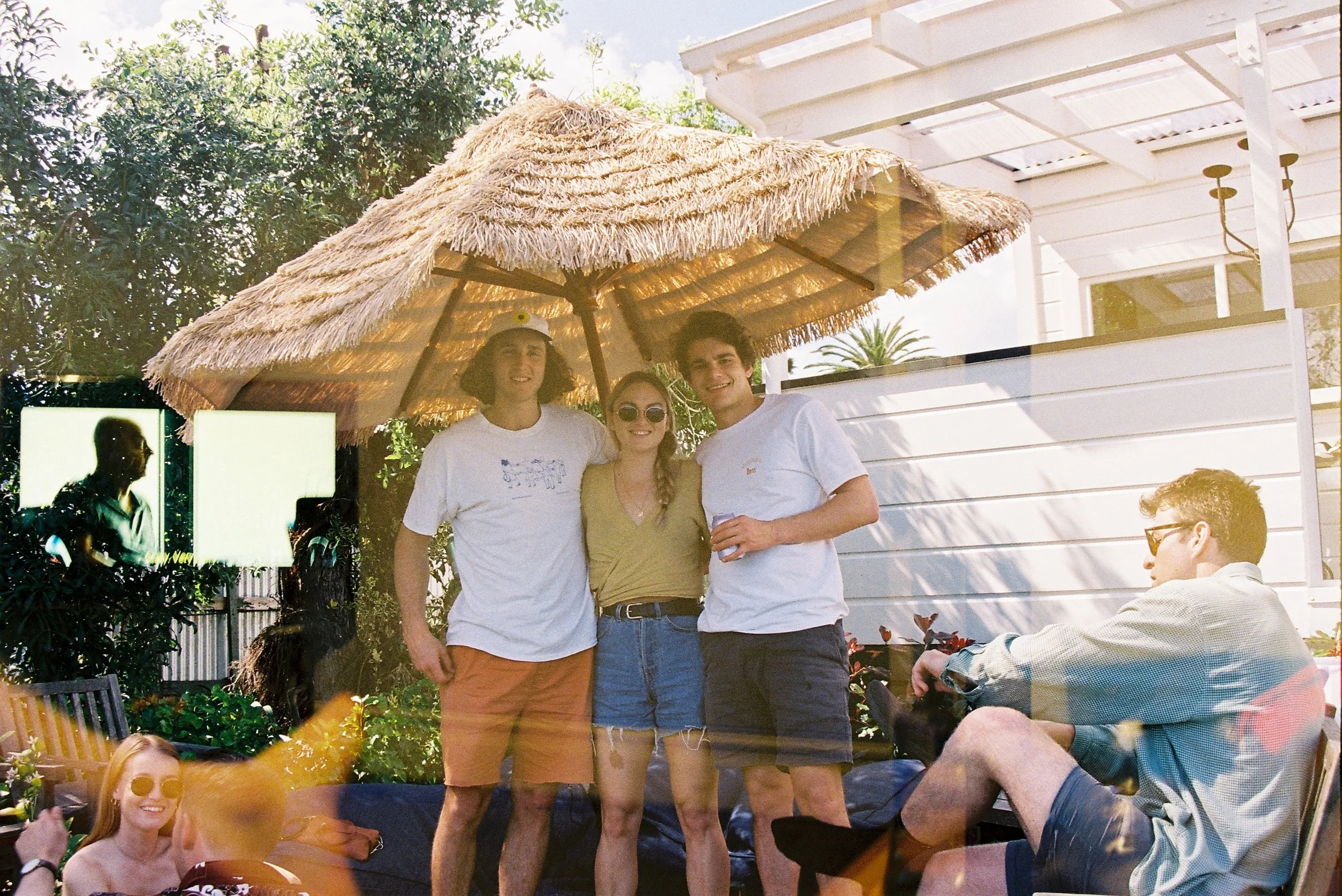 Group of four friends standing together outdoors under a thatched umbrella on a sunny day, with a white house in the background, some friends sitting and relaxing nearby, and trees around.
