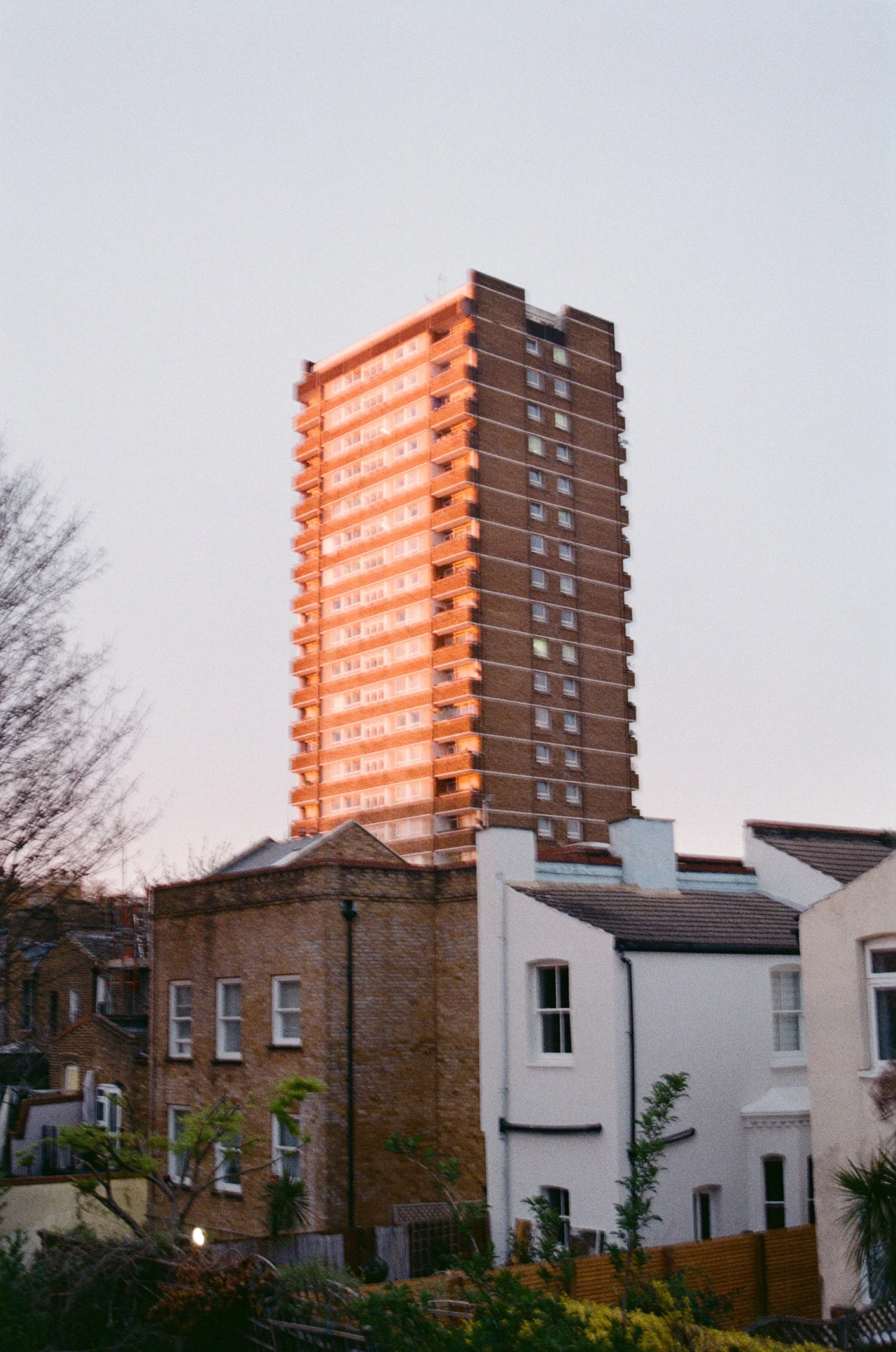 A tall residential building with multiple floors reflected in the sunset, with older houses and trees in the foreground.