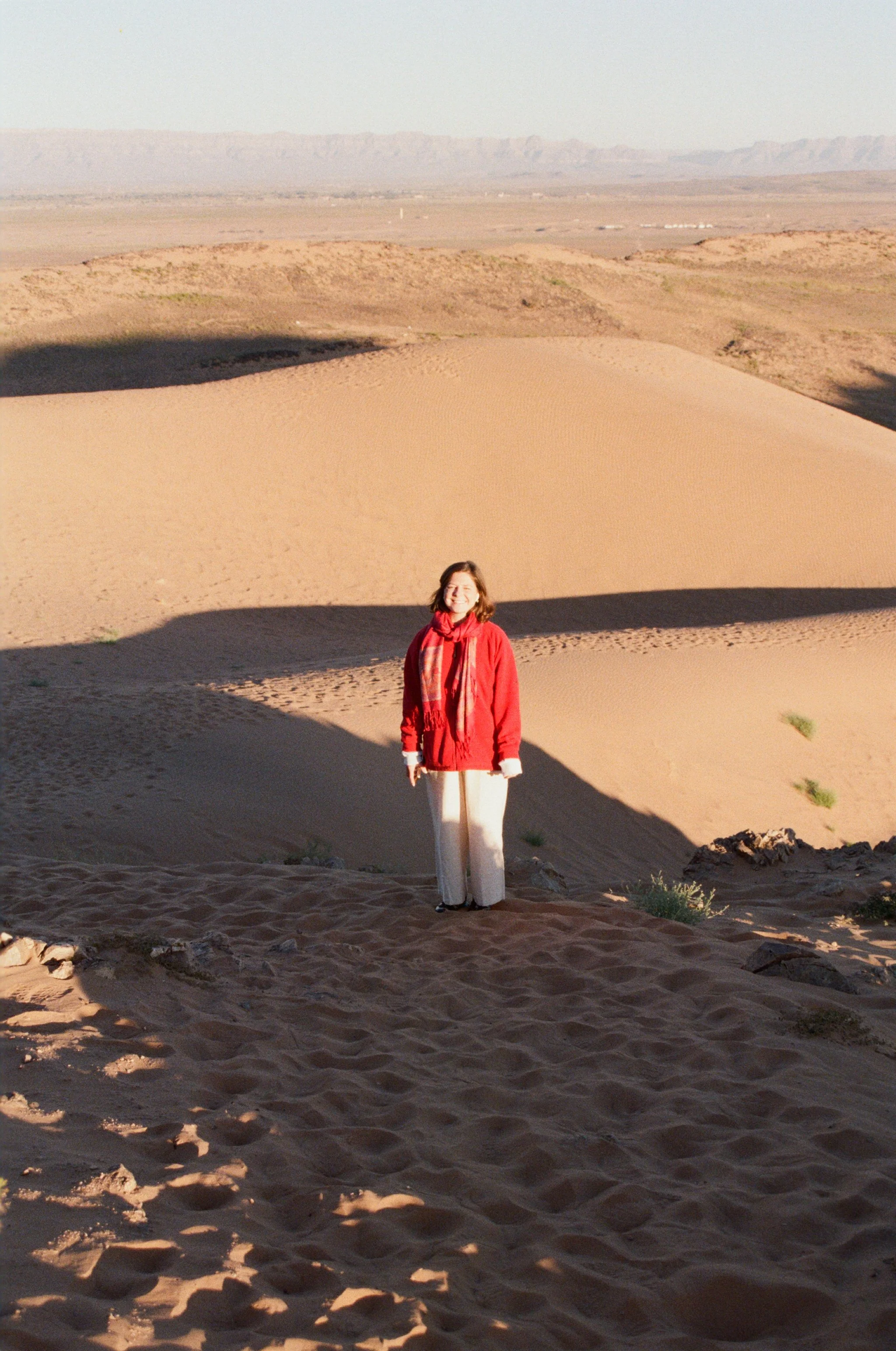 A woman standing in a sandy desert with sand dunes, wearing a red jacket and white pants, smiling at the camera, with mountains in the background.