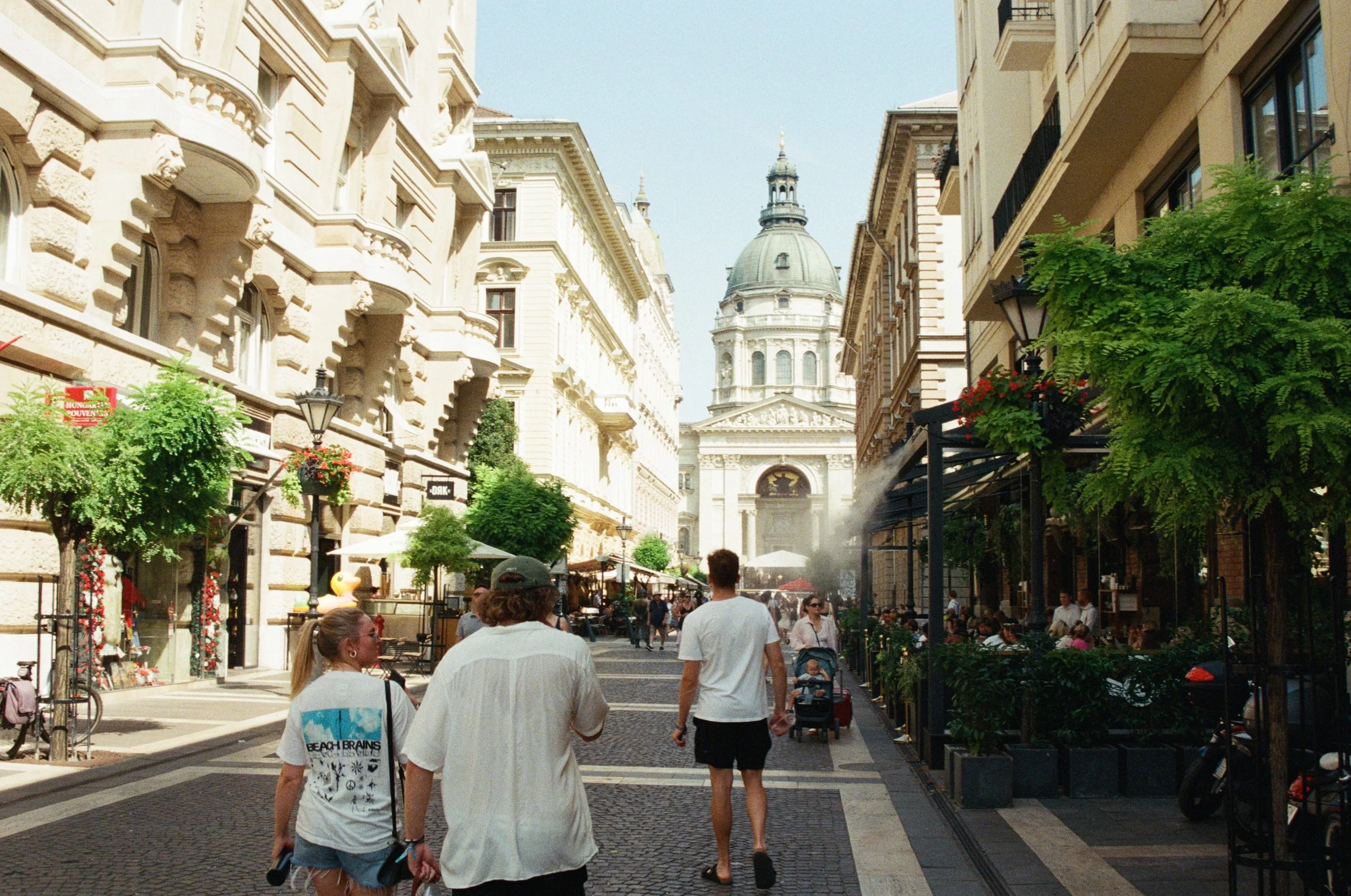 People walking down a city street with historic buildings and a domed church in the background.