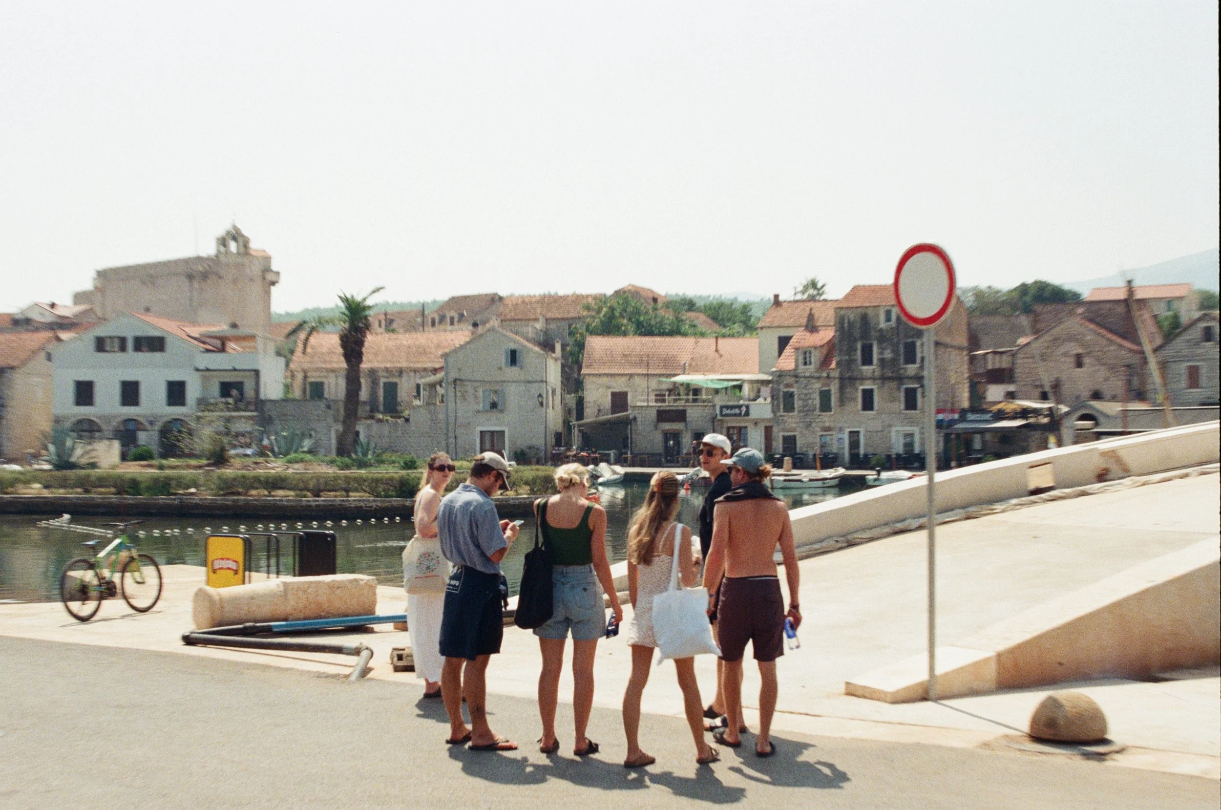 Group of people standing near a waterfront with traditional Mediterranean-style houses in the background, sunny day.