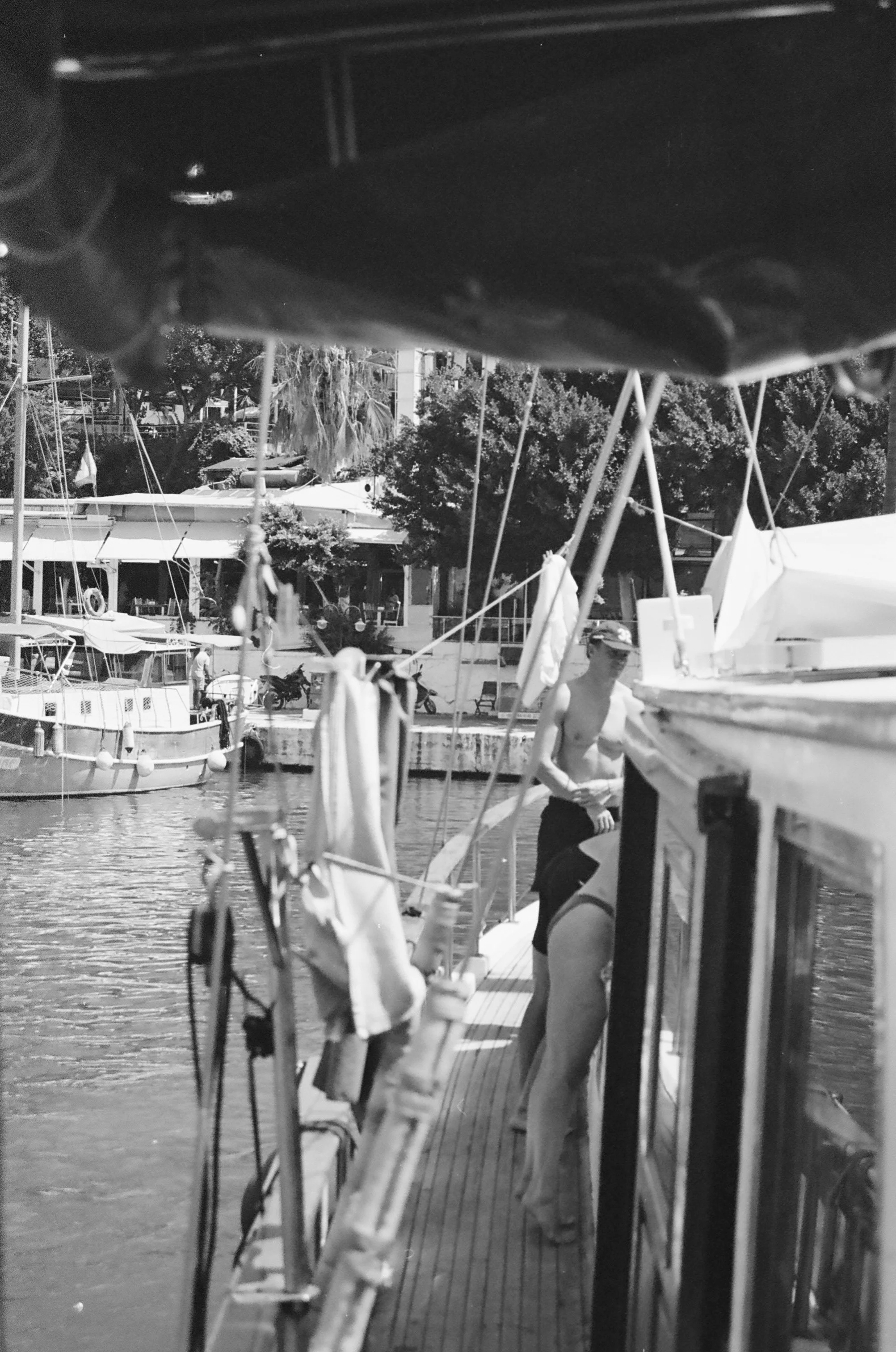 Black and white photo of a dock with several boats, two shirtless men are standing on the dock, one in the foreground leaning on the boat and the other in the background facing sideways, with trees and a building in the background.