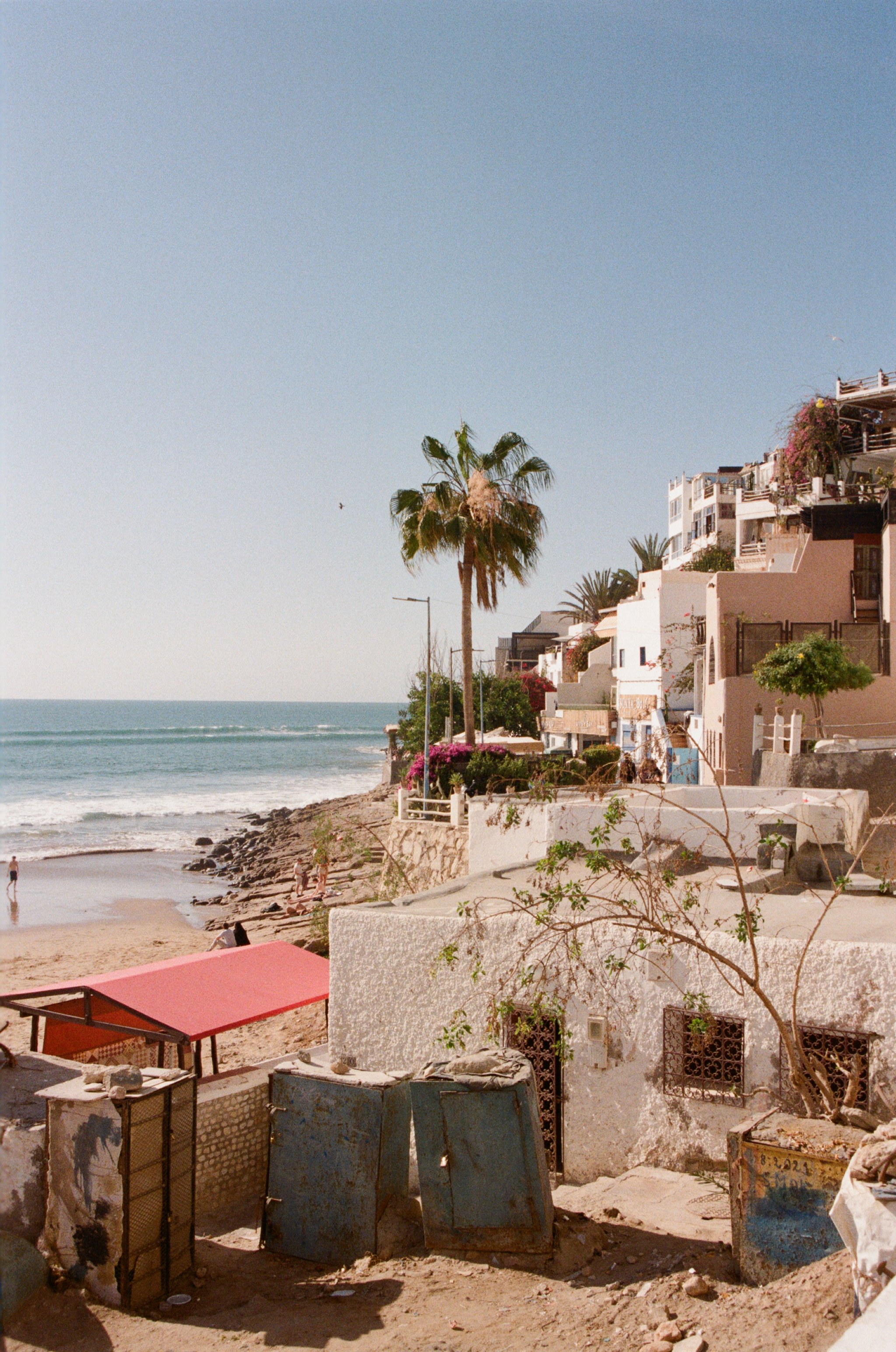 Beachside scene with a sandy beach, ocean waves, palm trees, and colorful houses on a hillside.