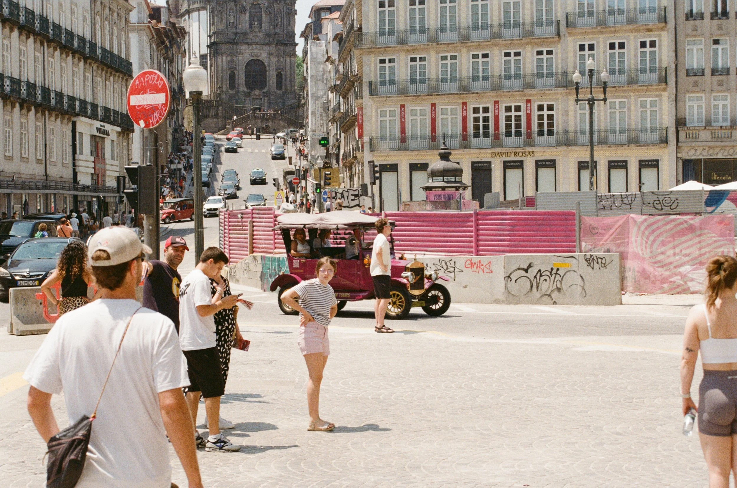 People waiting to cross a city street near a pink sightseeing vehicle, with buildings, parked cars, and graffiti in the background on a sunny day.