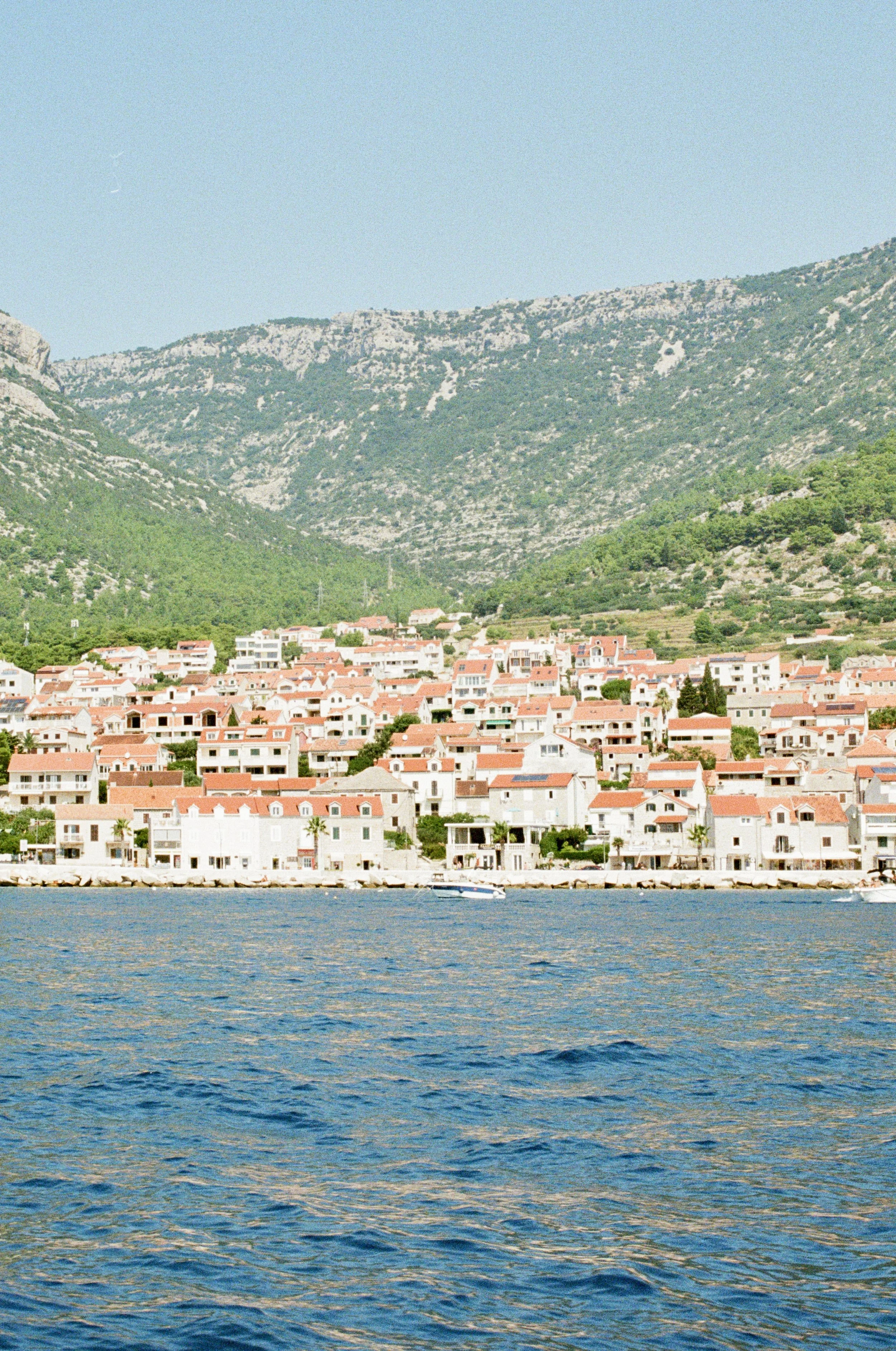 Coastal town with white buildings and red roofs along the shoreline, with green mountains in the background under a clear blue sky.
