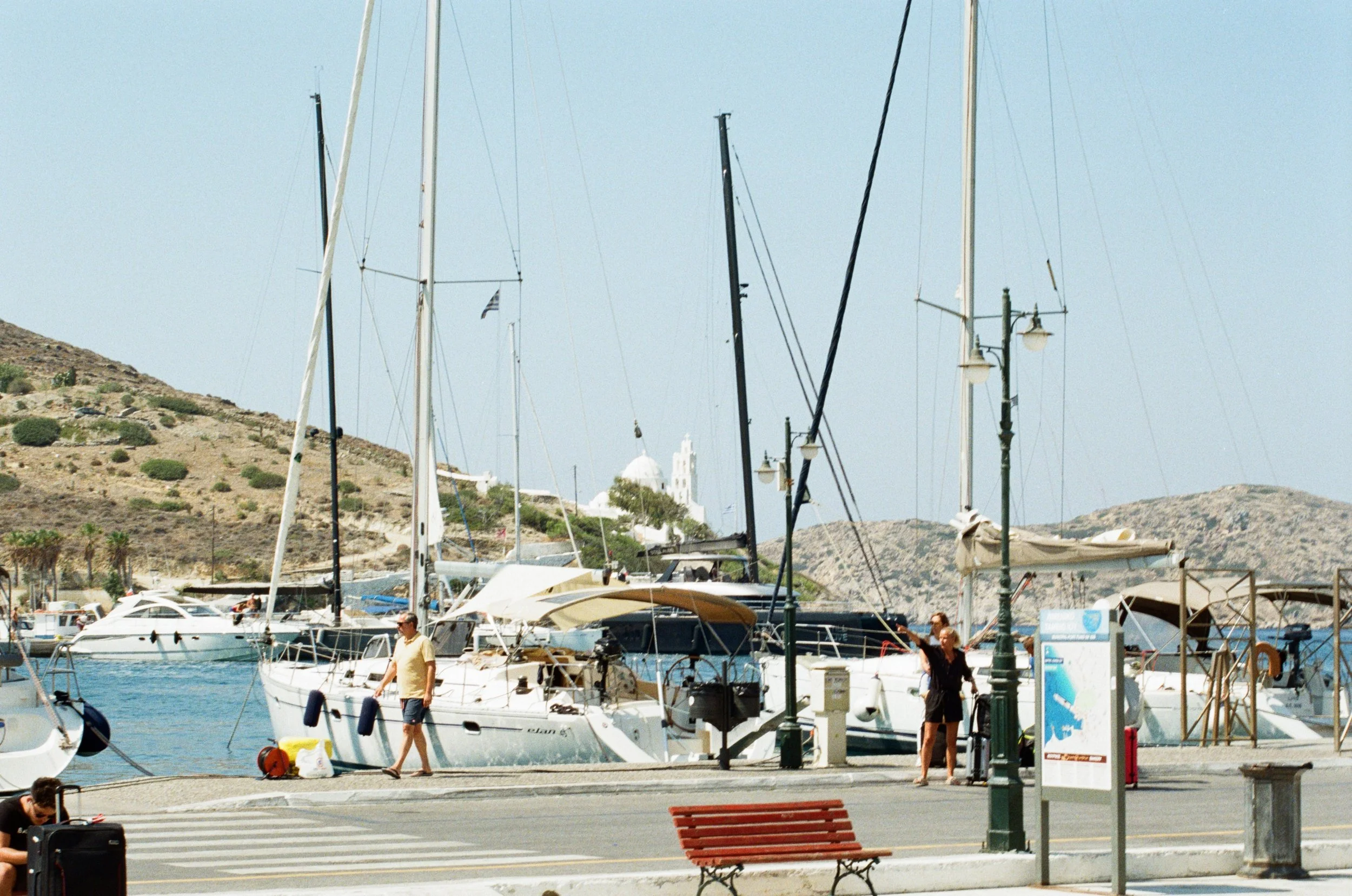 People walking along a dock by a marina with sailboats and yachts, hillside with sparse vegetation in the background.