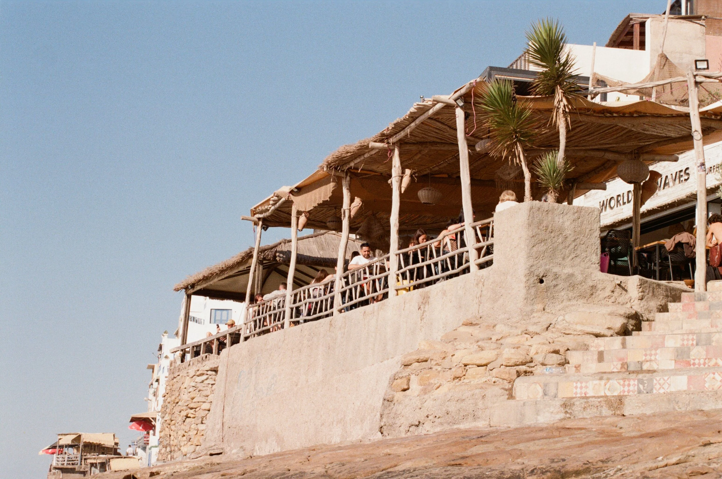 People dining outdoors under a thatched roof at a seaside restaurant with a stone and concrete deck, palm trees, and a clear blue sky.