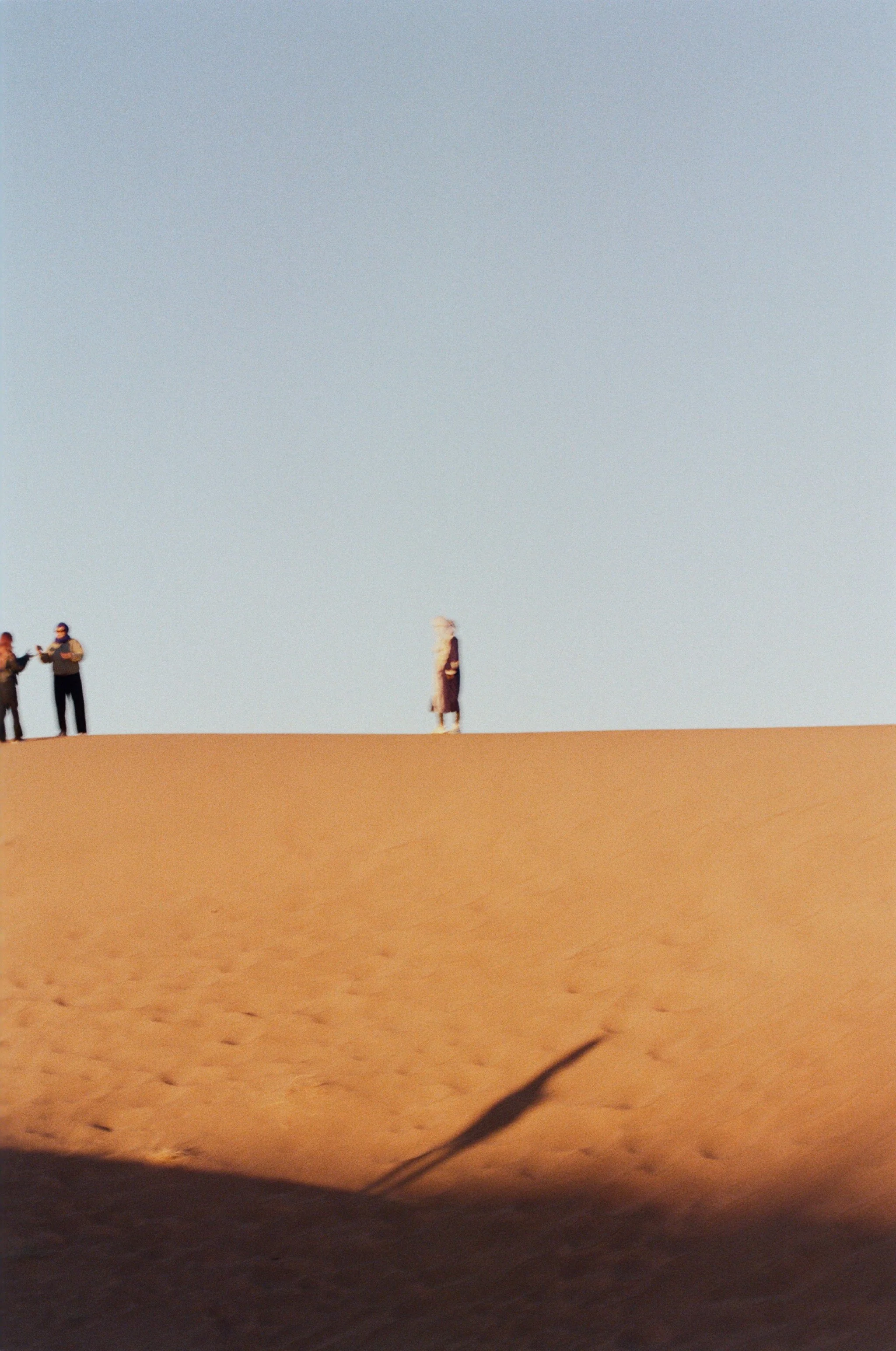 A desert landscape with a few people standing on the sand dunes against a clear sky.