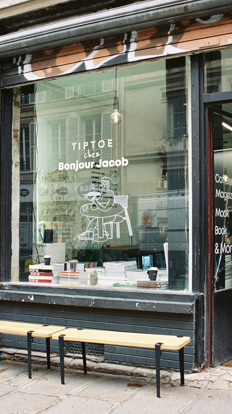 Store window with white line art of a person reading a book at a table, surrounded by books and a coffee cup. Text on the window says "TIPTOE chez Bonjour Jacob."