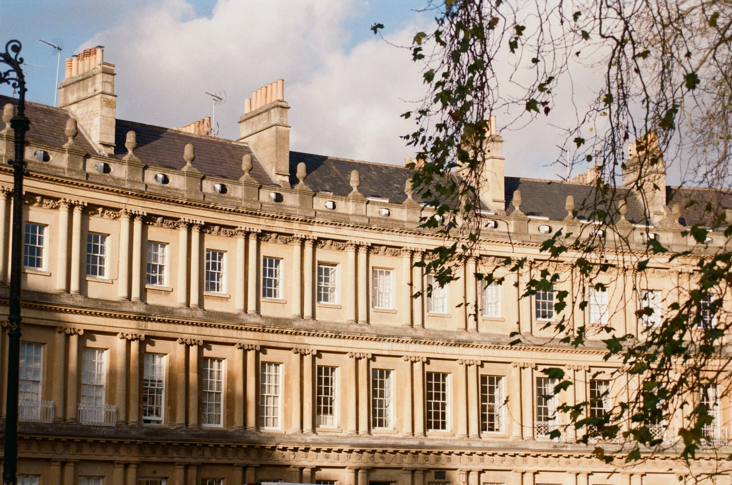 A historic building with classic architecture, multiple stories, beige stone exterior, numerous windows, and chimney stacks on the roof. Leafy branches partially cover the view.