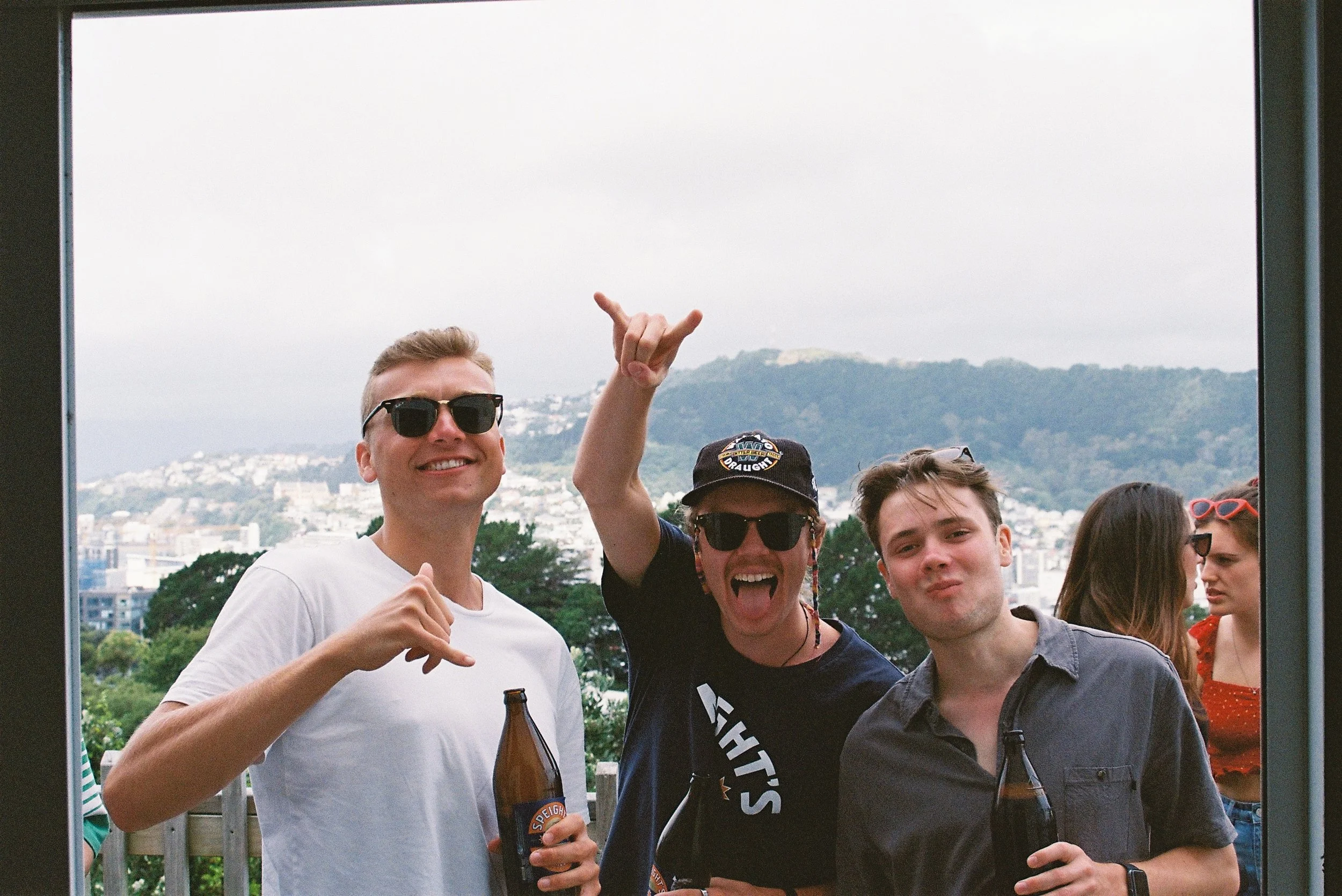 Three young men outdoors celebrating with drinks, smiling and making gestures, with a cityscape and hills in the background, during daytime.