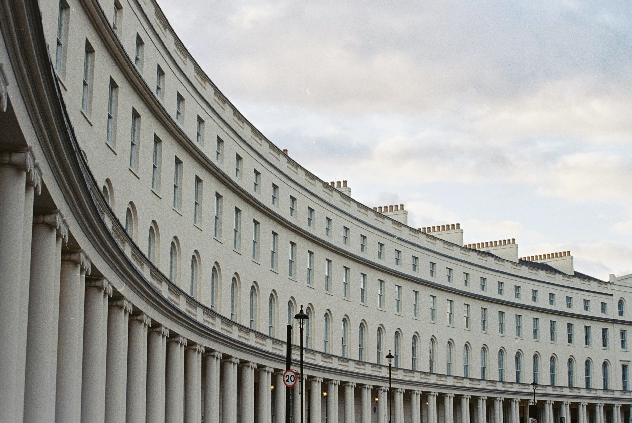 White curved historical building with multiple windows and columns, under a cloudy sky, with streetlights and a 20 mph speed limit sign.