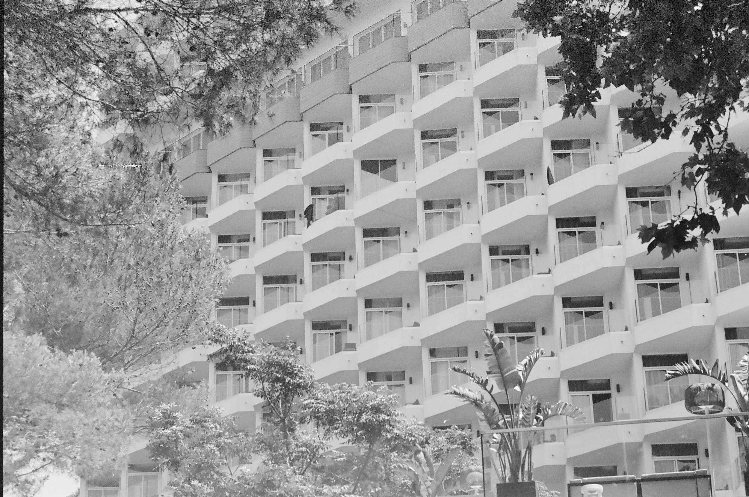 A multi-story apartment building with repeating balconies, surrounded by trees and plants.