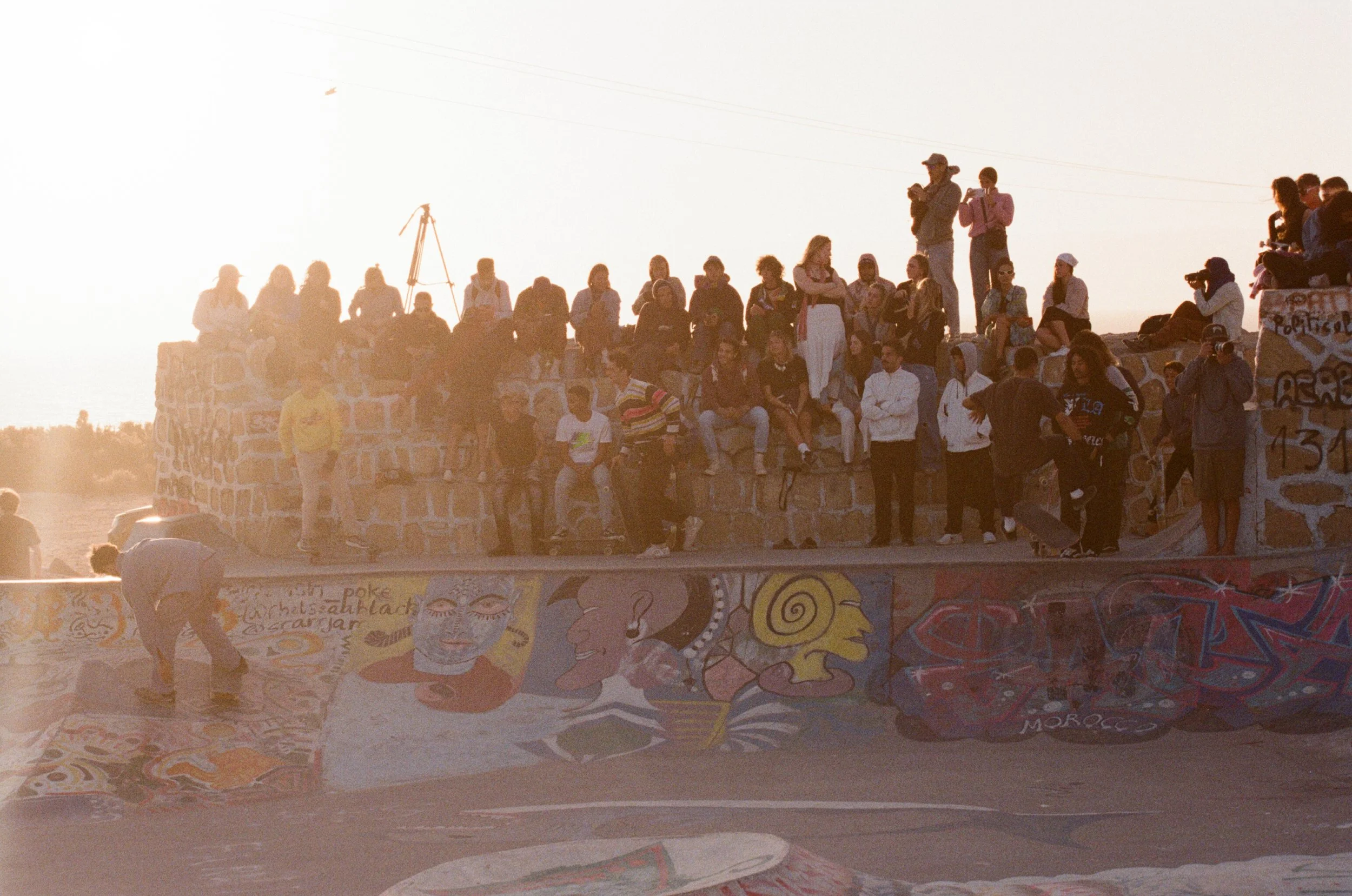 A large group of people sitting and standing on a stone skatepark ledge, with colorful graffiti art on the ramp below, during sunset.