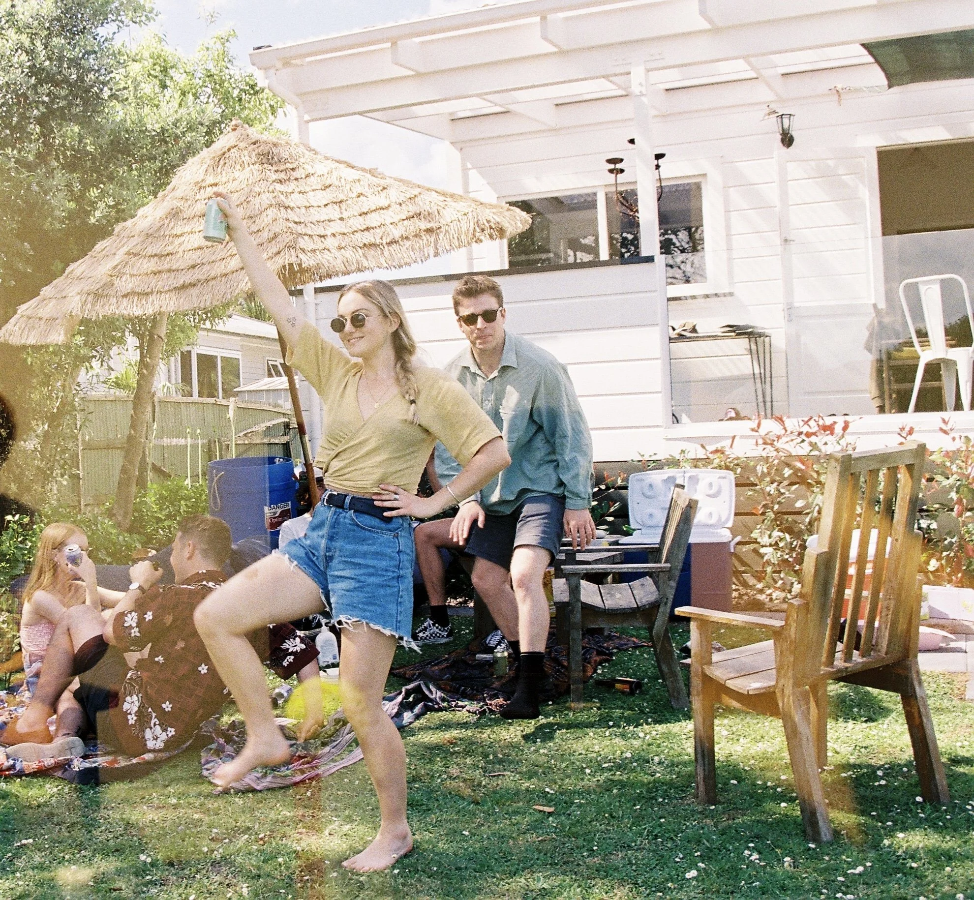 A group of people enjoying a backyard gathering on a sunny day, with a woman dancing in front, holding a drink, wearing sunglasses, a yellow shirt, and blue denim shorts. In the background, others are sitting on the grass, talking, and drinking near 