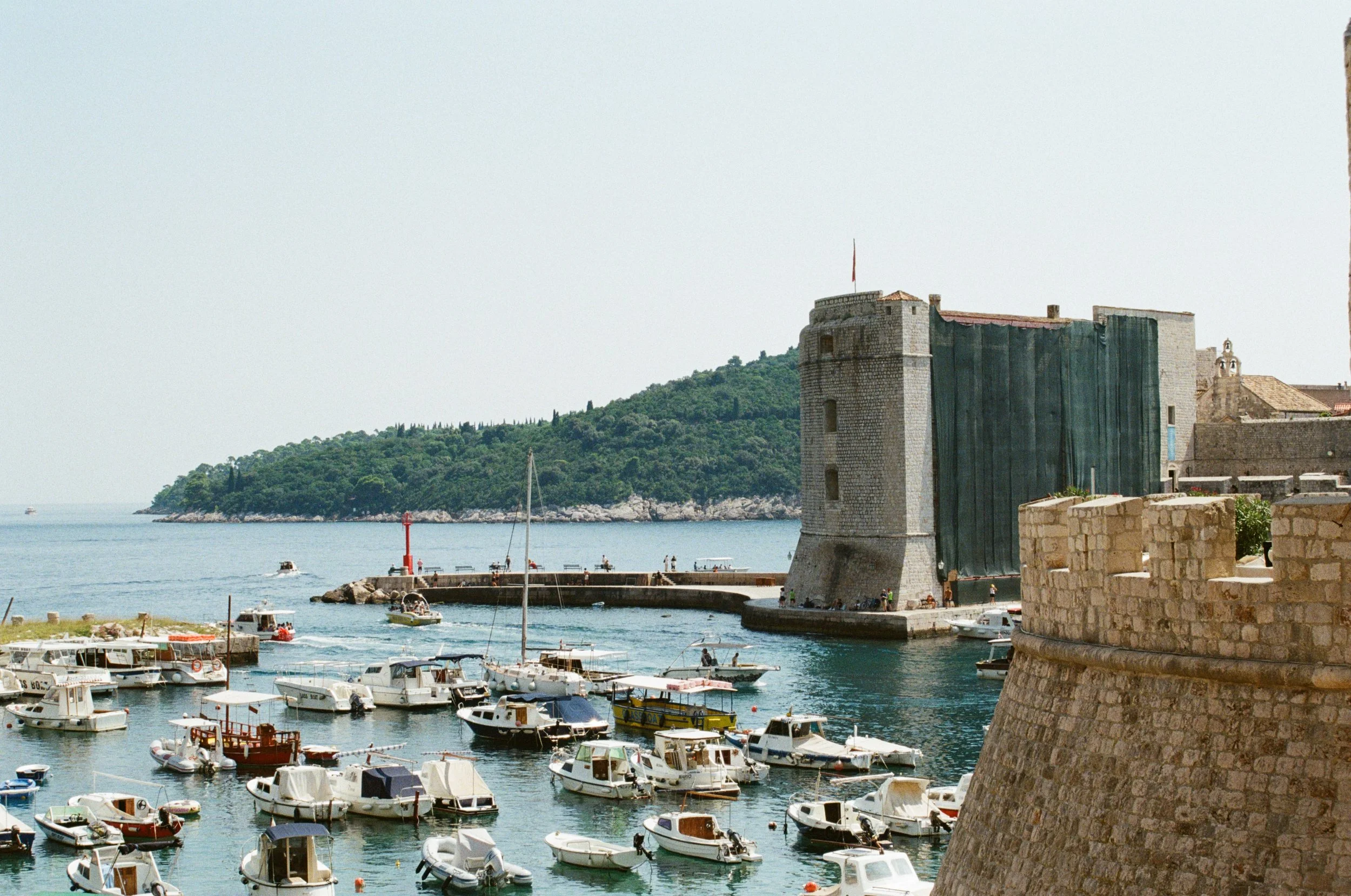 A harbor with numerous boats docked, an old stone fortress with a green curtain hanging from it, and a lush green island in the background.