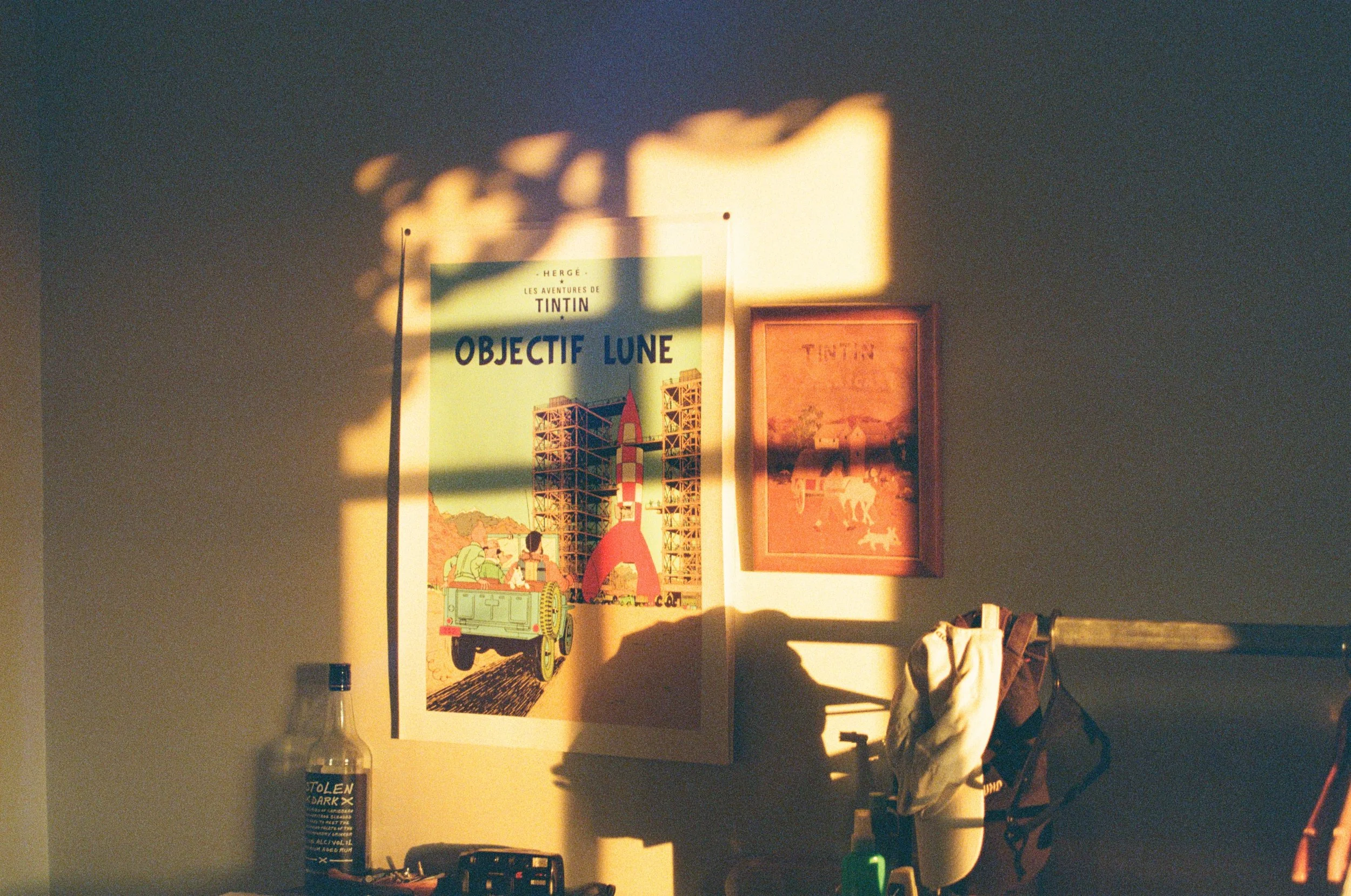 Sunlight casting shadows on a wall with framed art, including a poster of a rocket and cityscape, along with various personal items and clothing.