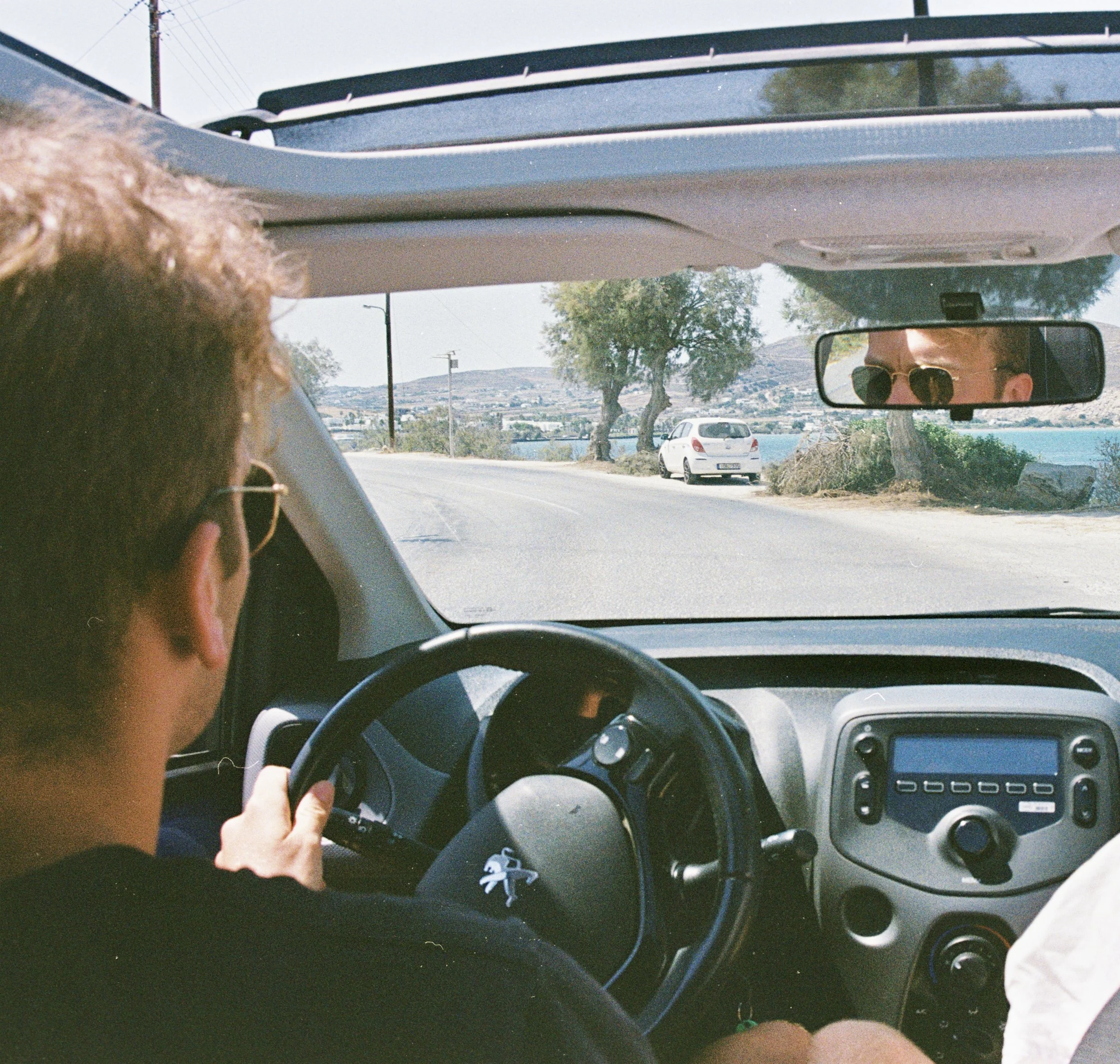Interior view of a car with a person driving, looking at the road ahead, with a rearview mirror reflecting another person's face with sunglasses, and a scenic view of a road, trees, and water outside.