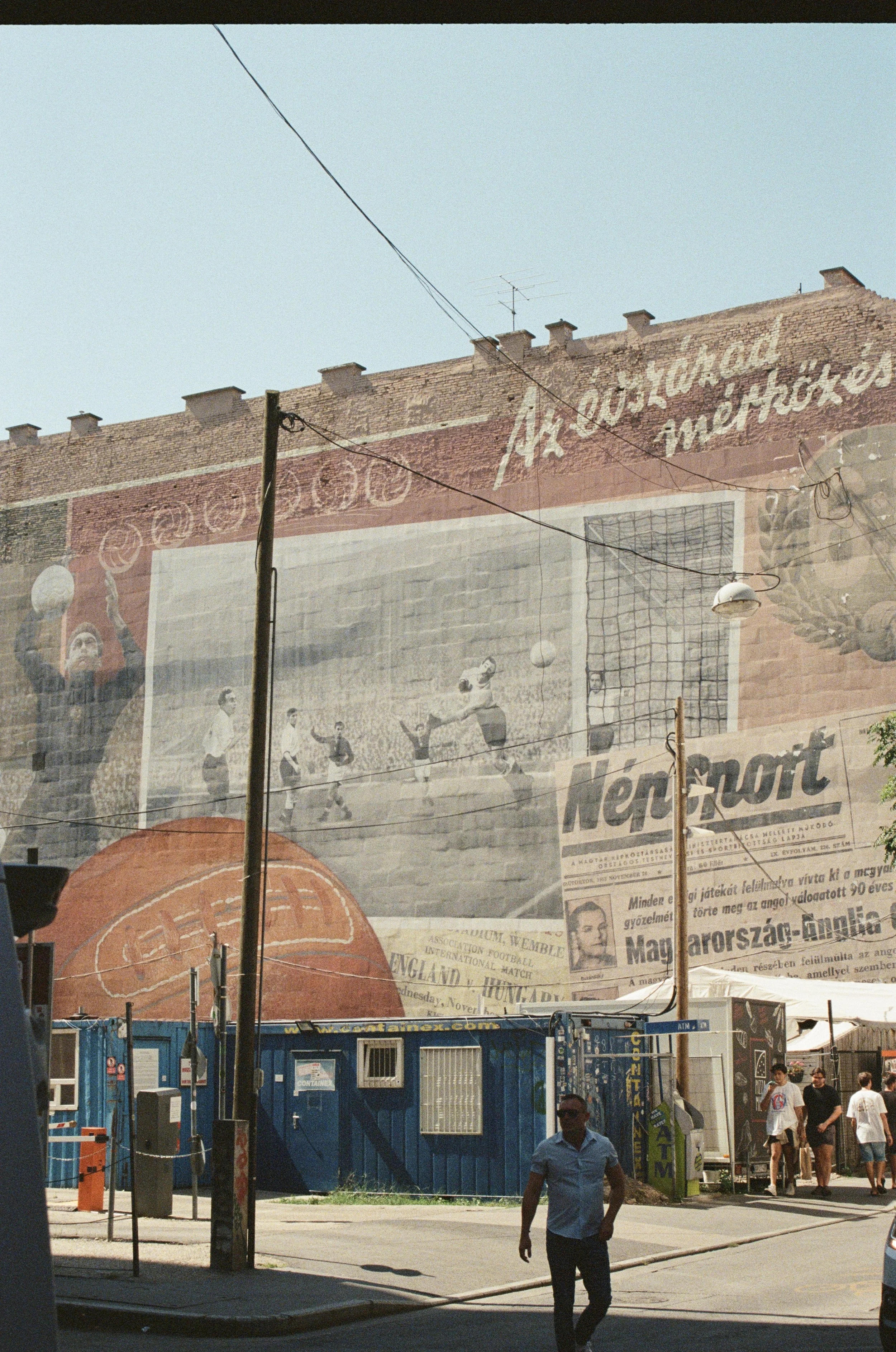 A large mural painted on the side of a brick building, depicting vintage sports scenes, including basketball, soccer, and a newspaper headline about a Hungarian football match.