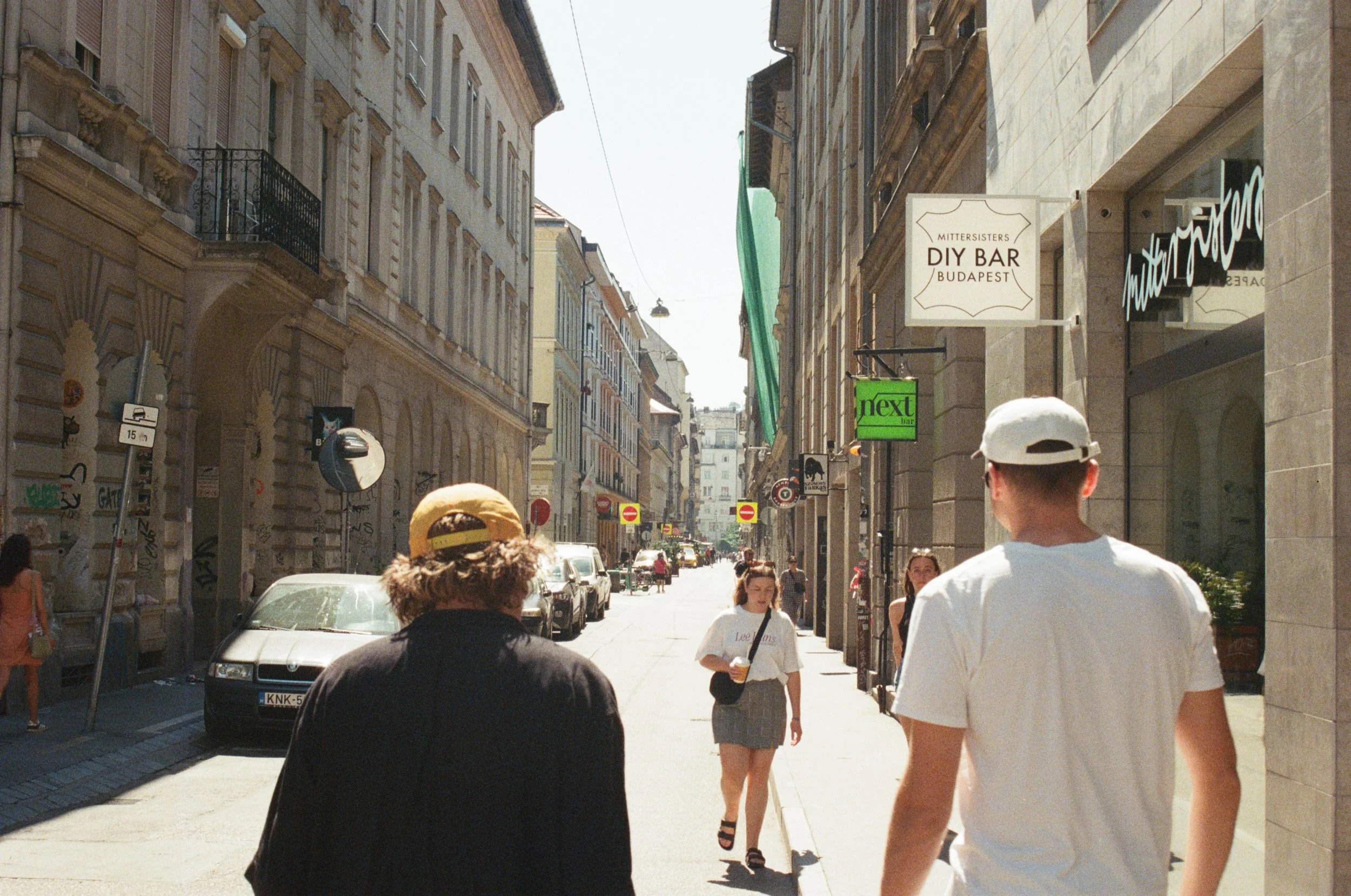 Street scene in Budapest featuring pedestrians walking along a sunny sidewalk, with buildings on either side and signs for local bars and businesses visible.