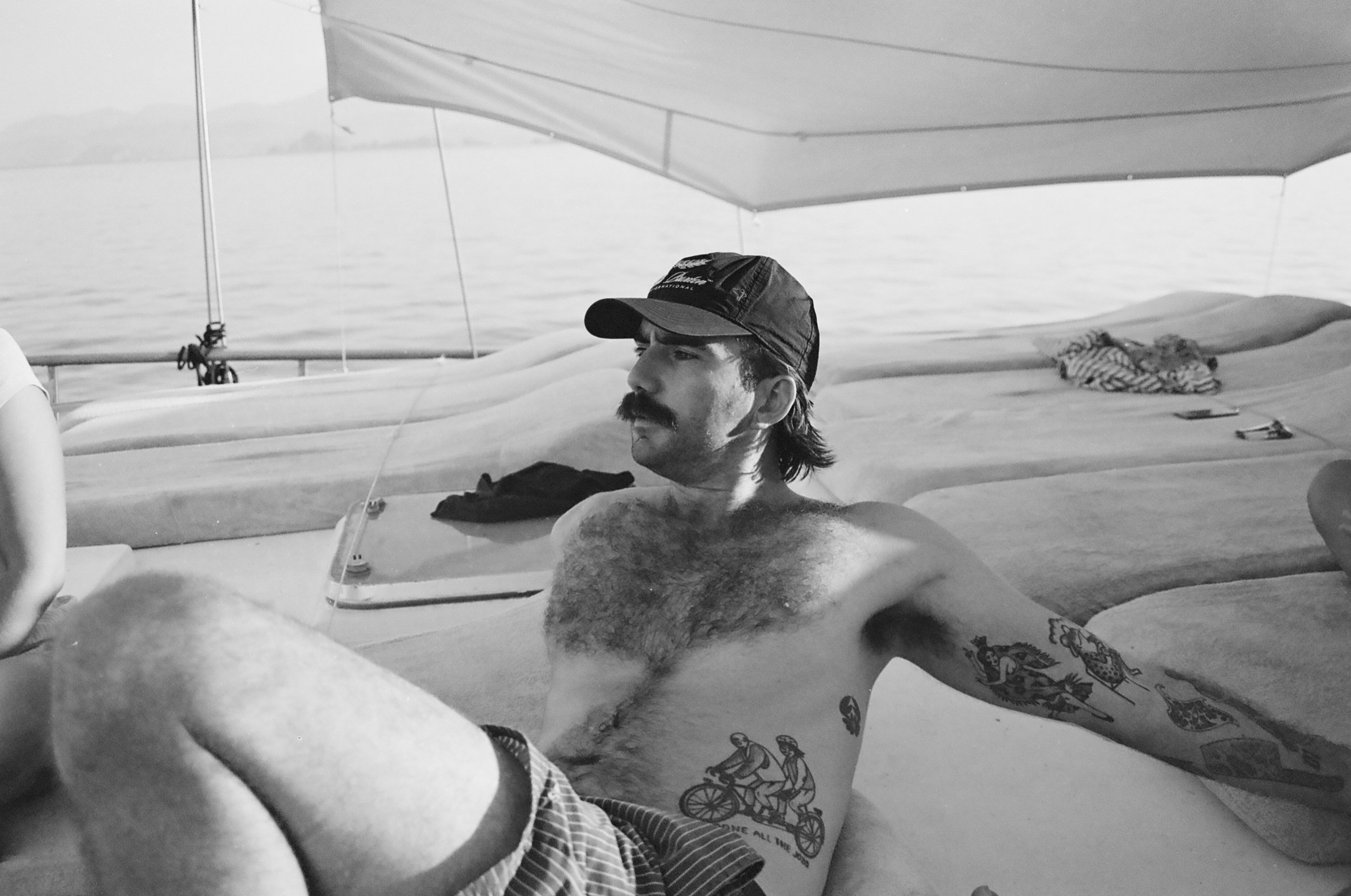 A shirtless man with a mustache and tattoos, wearing a cap, sitting on a boat with water in the background and a canopy above.