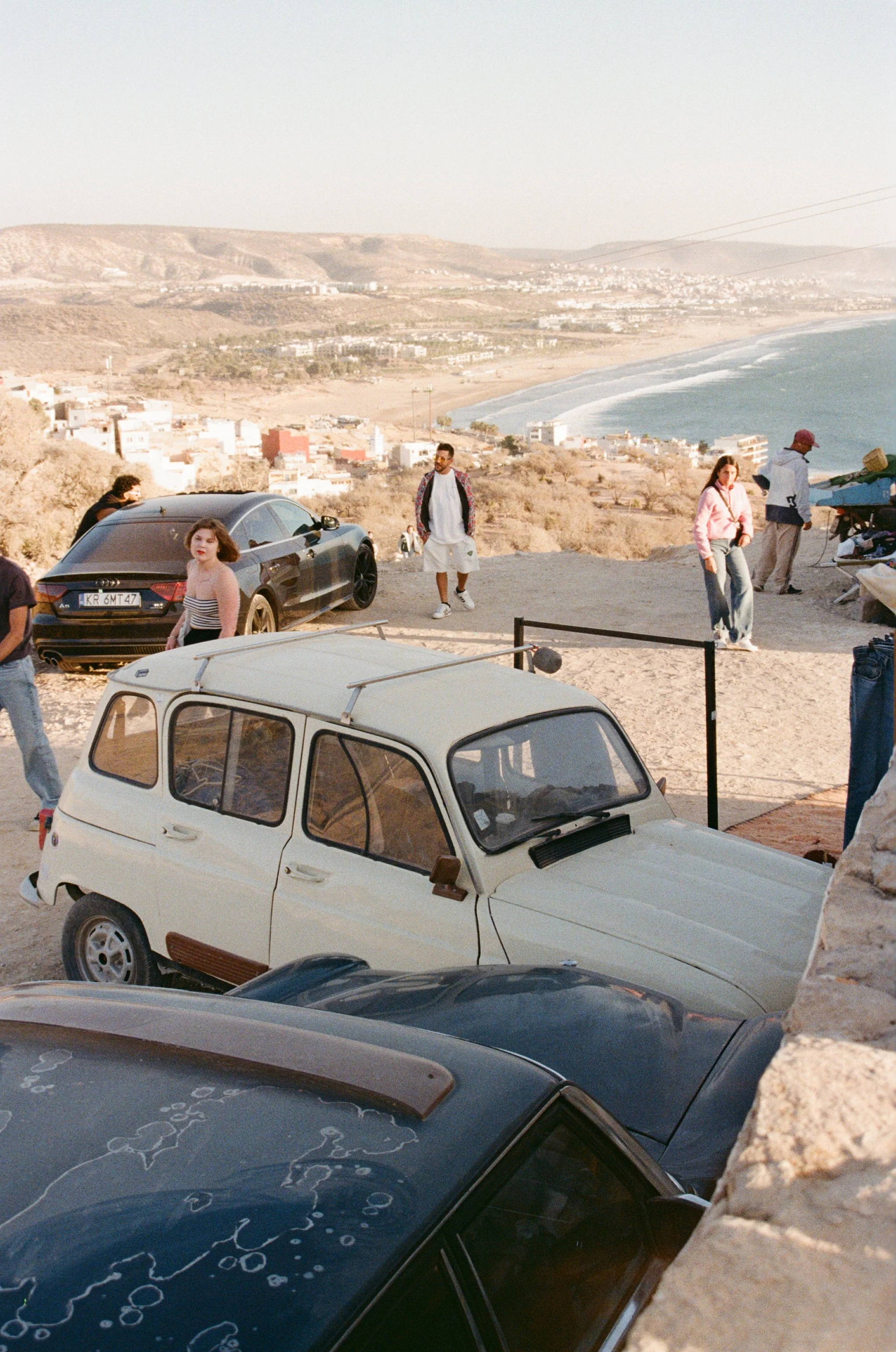 A coastal scene with a parking area on a hill overlooking the ocean, featuring several parked cars and people walking, with a view of a beach, hills, and a distant town in the background.