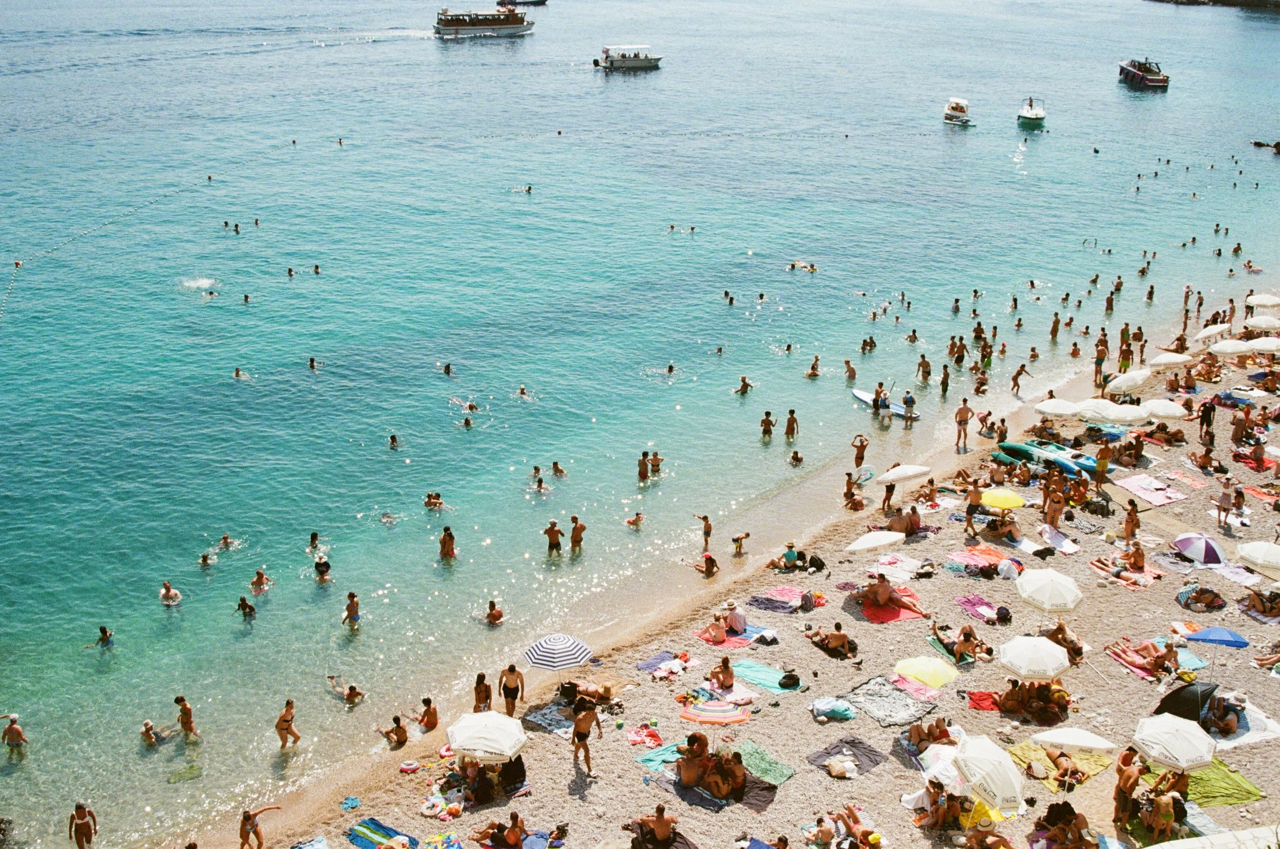 Beach scene with many people swimming, sunbathing, and relaxing on colorful towels and umbrellas along a sandy shore. Several boats are anchored offshore in the calm, clear water.