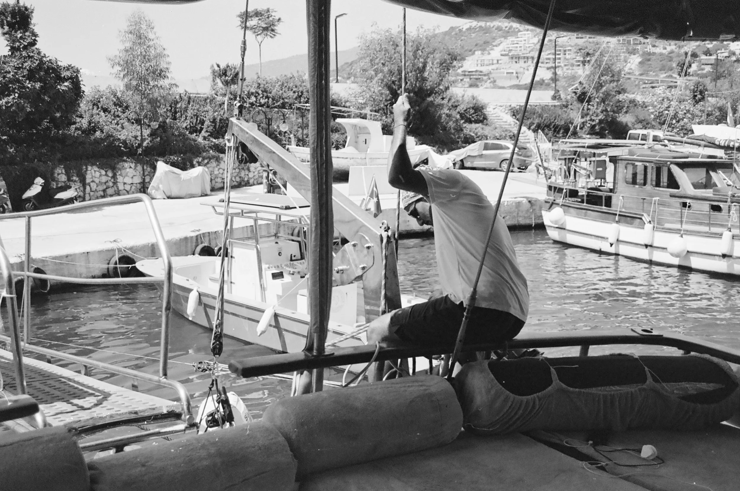 A man in a t-shirt and cap working on a small sailboat at a marina, with other boats and boats covered with tarps in the background.
