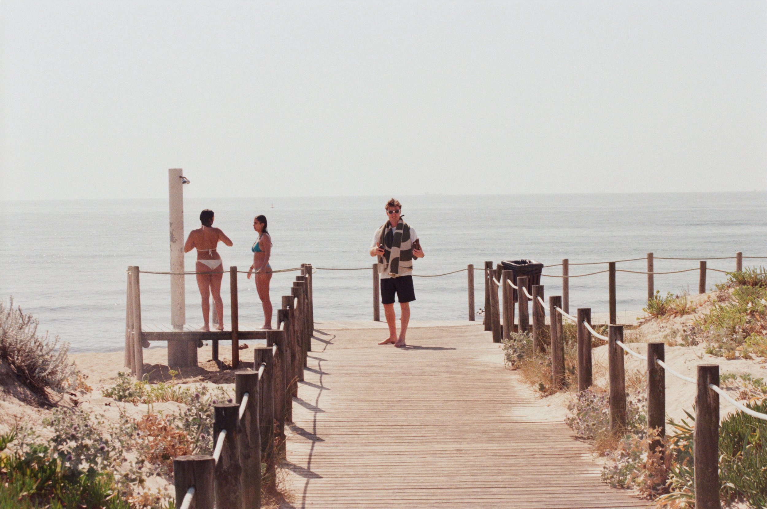A person walking along a wooden boardwalk towards the beach, with two women in swimsuits near a shower station and the ocean in the background.