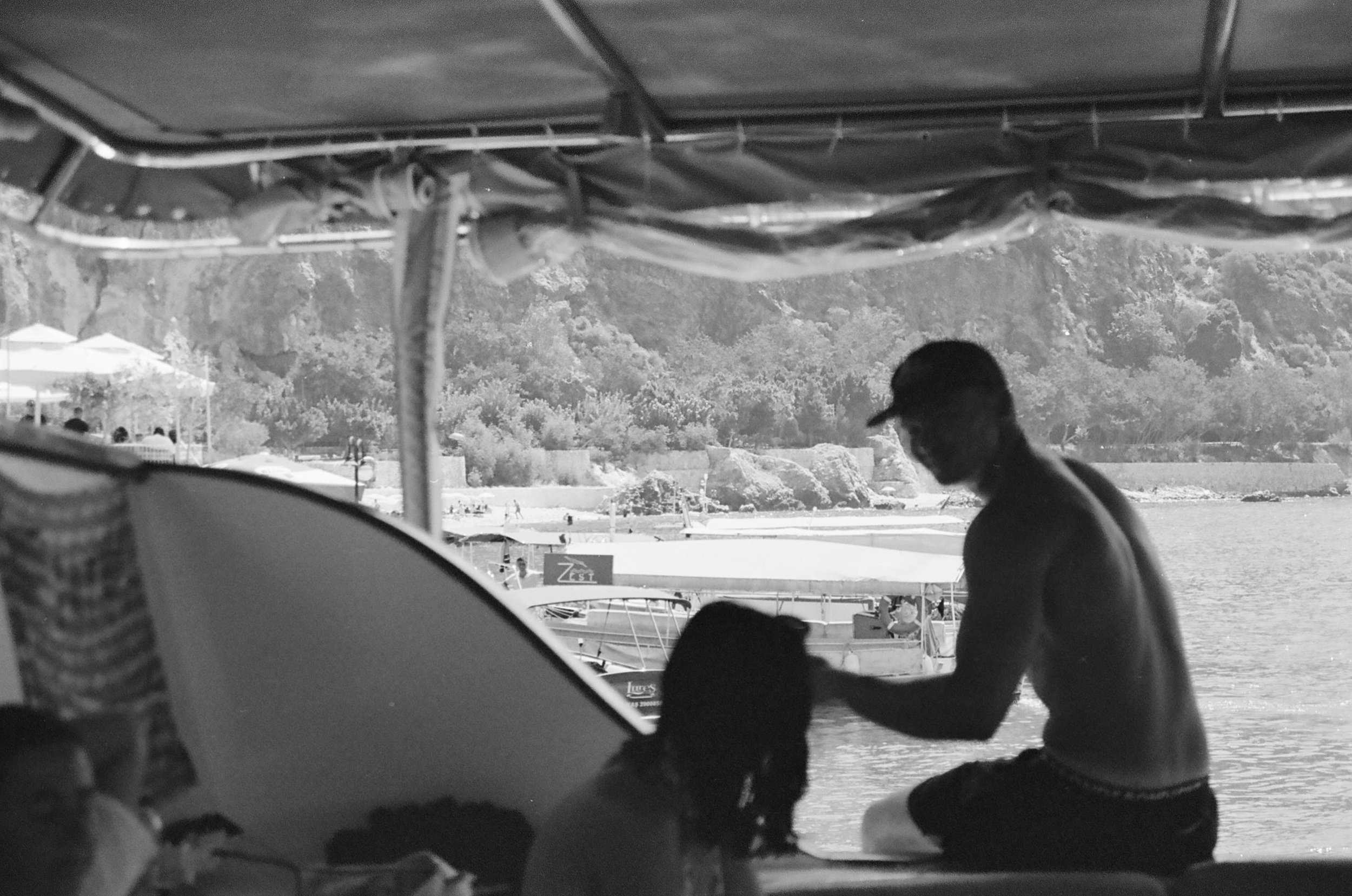 A black and white photo of a man and woman by the water, sitting under a canopy. The man is shirtless and wearing a cap, while the woman has long hair. There are boats and a shoreline visible in the background.