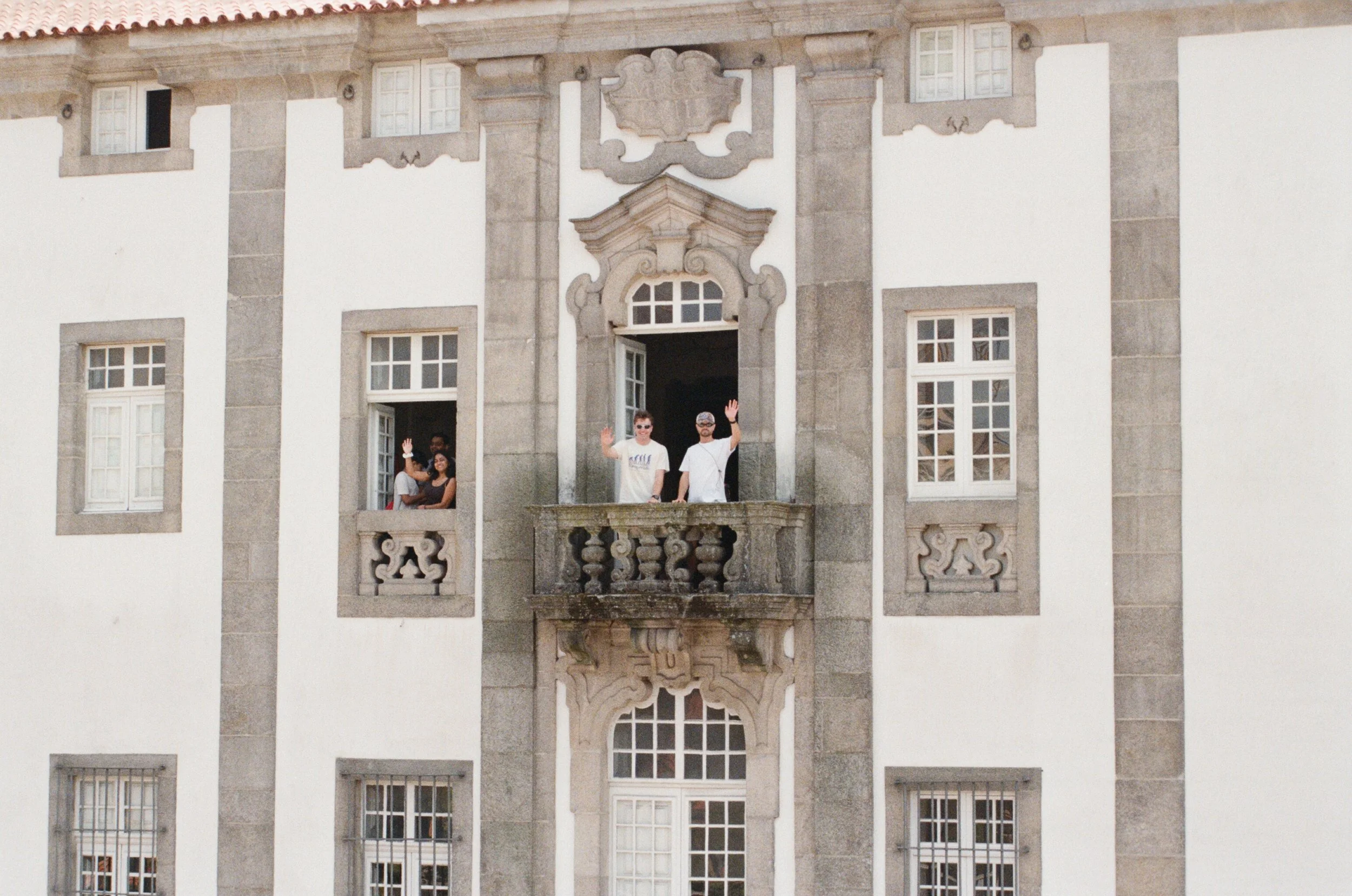 Three people standing on a balcony waving, with two others sitting at an open window, on the facade of a historic building with ornate architectural details.