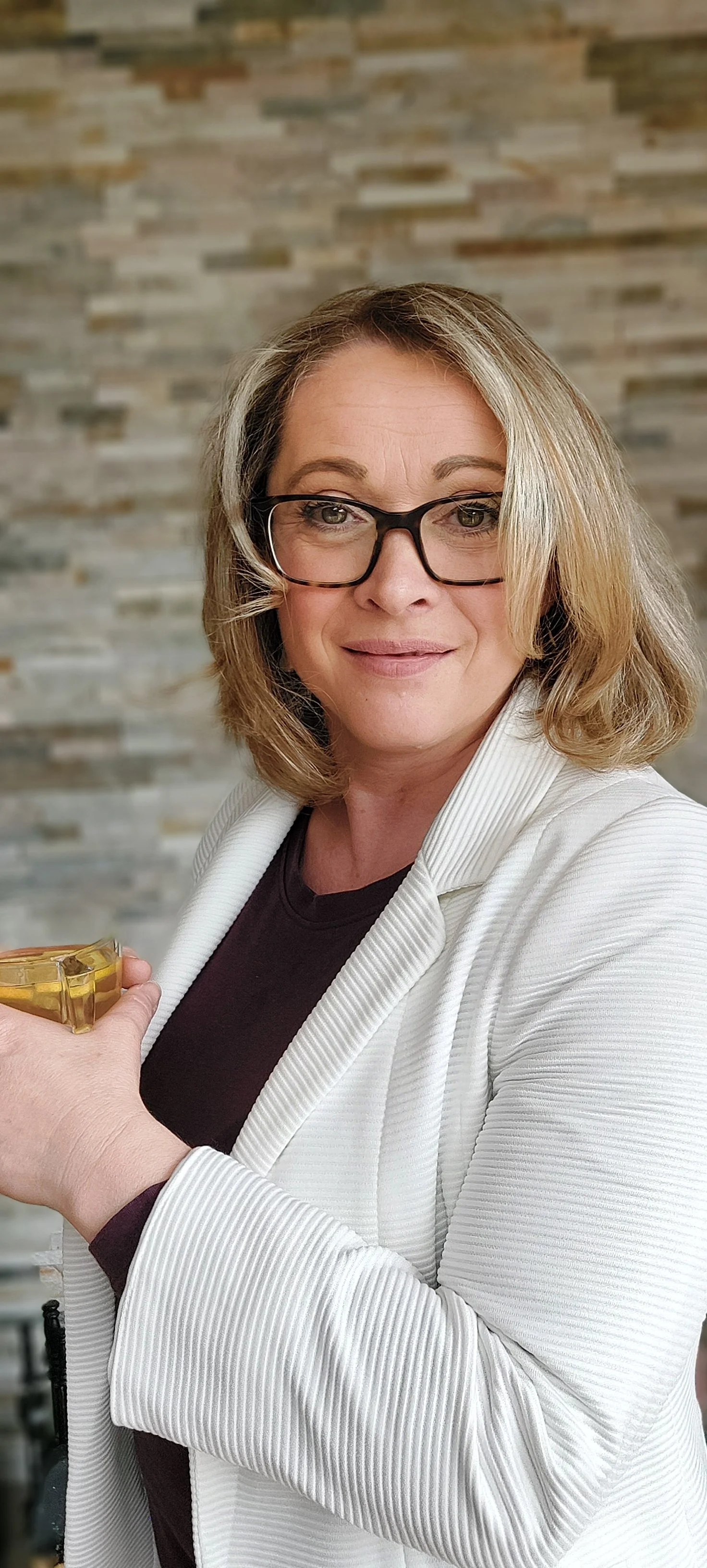 A woman with blonde hair, wearing glasses, a white blazer, and a black top, holding a yellow object, standing in front of a stone wall.