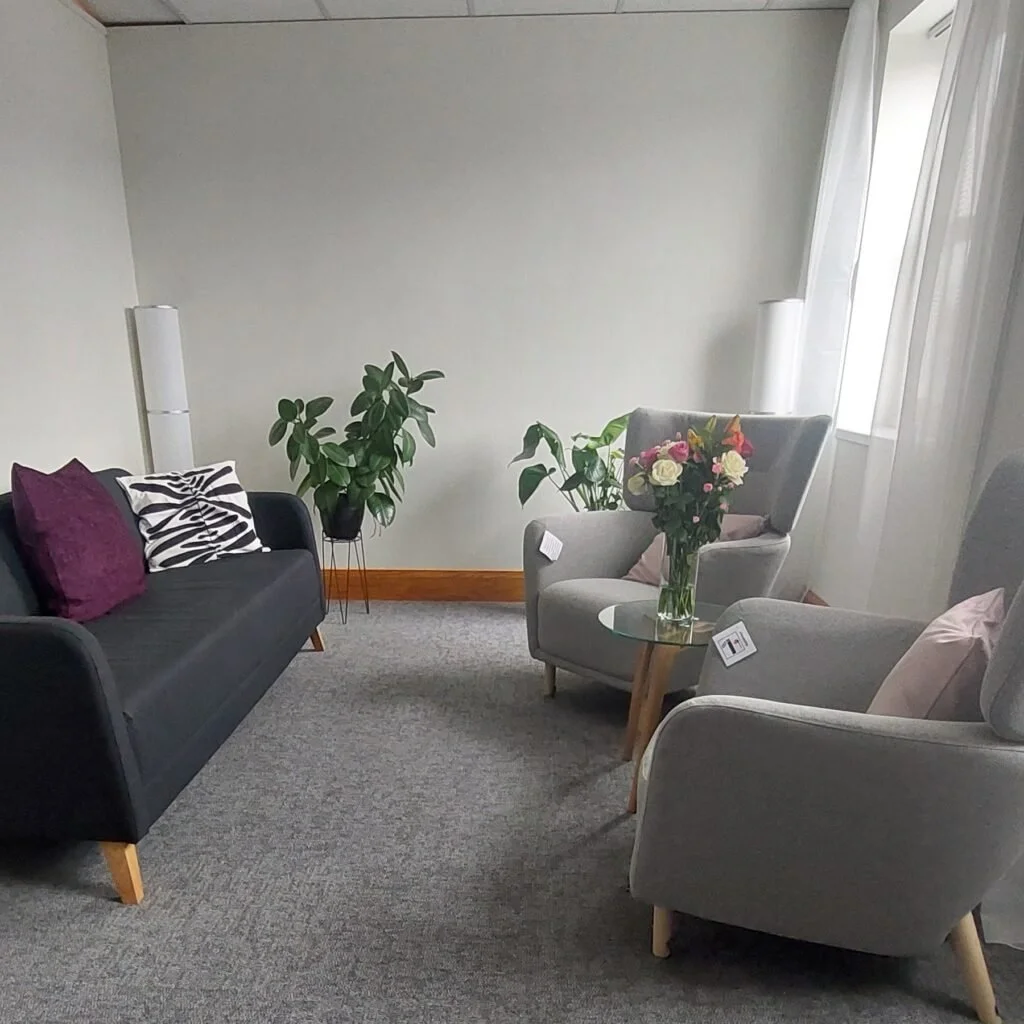 Living room with a gray sofa, two gray armchairs, and a glass top side table holding a large bouquet of mixed roses. There are plants in the background, a corner window with sheer white curtains, and minimalist white lamps with tall speakers.