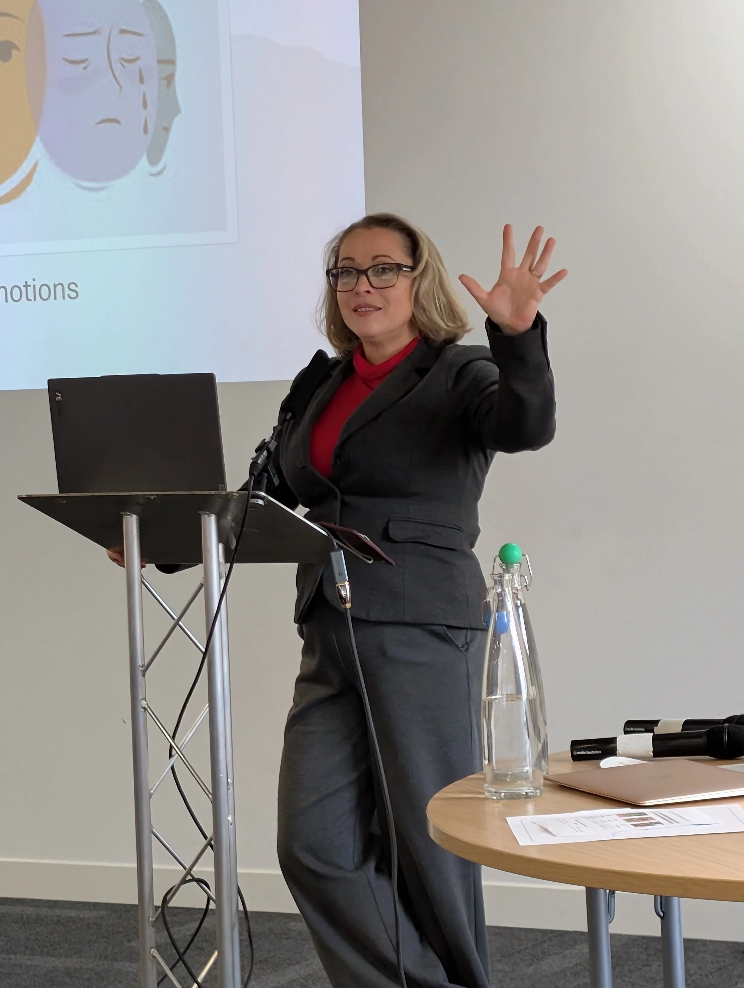 Woman giving a presentation, standing at a podium with a laptop, gesturing with her hand, and a projection of a slide behind her.