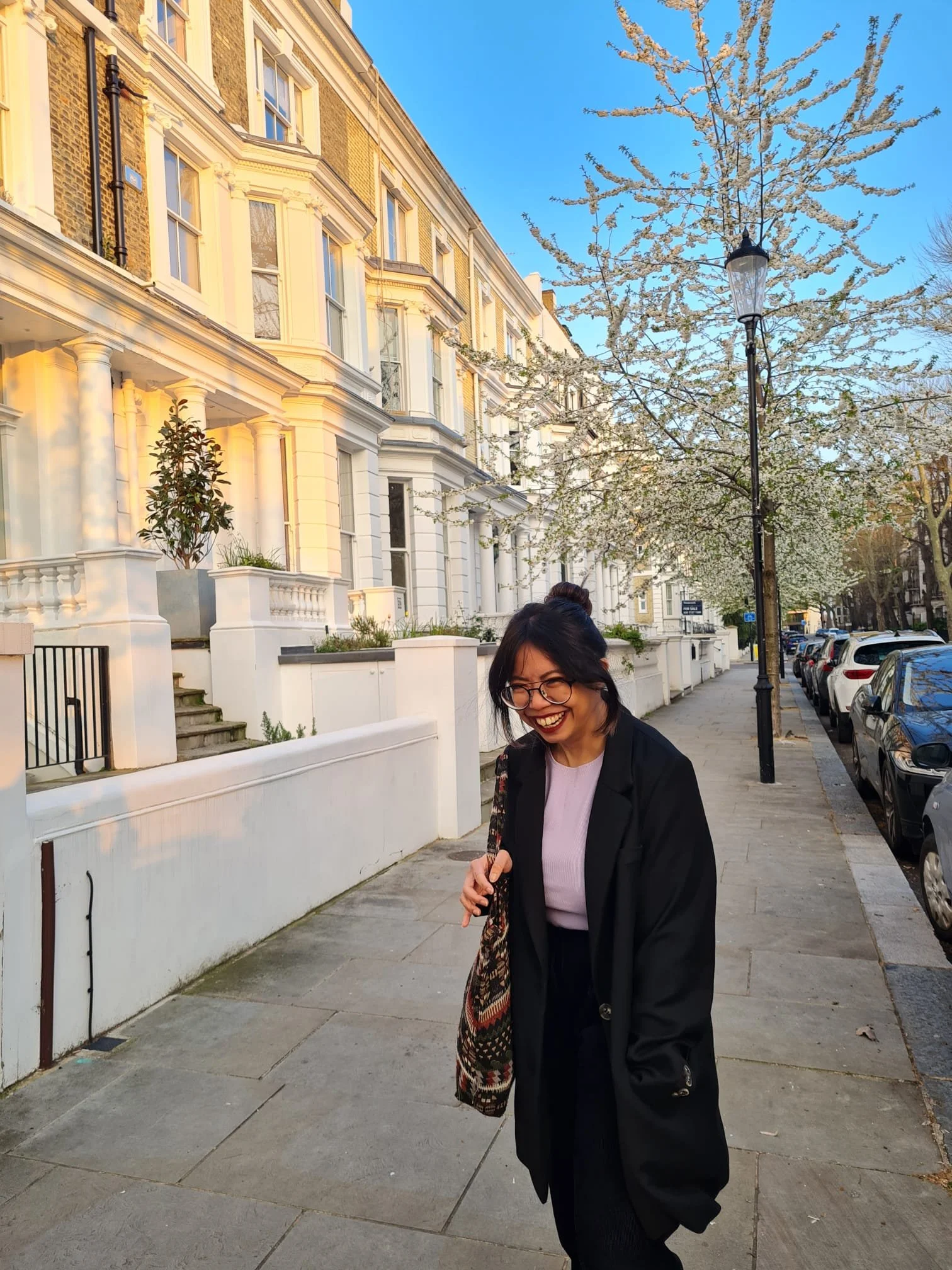 A woman with glasses and black hair smiling as she walks down a sidewalk in front of white row house buildings during springtime with blooming trees and parked cars on the street.