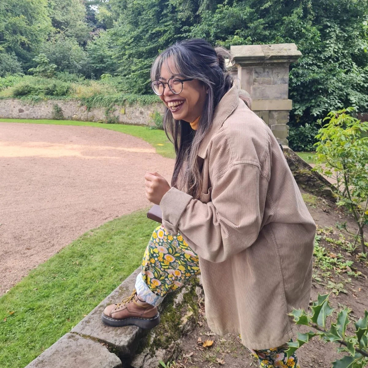 A woman with glasses and gray hair, wearing a beige jacket and floral pants, is sitting on a stone wall outdoors, laughing with a joyful expression. There is greenery and a gravel area in the background.