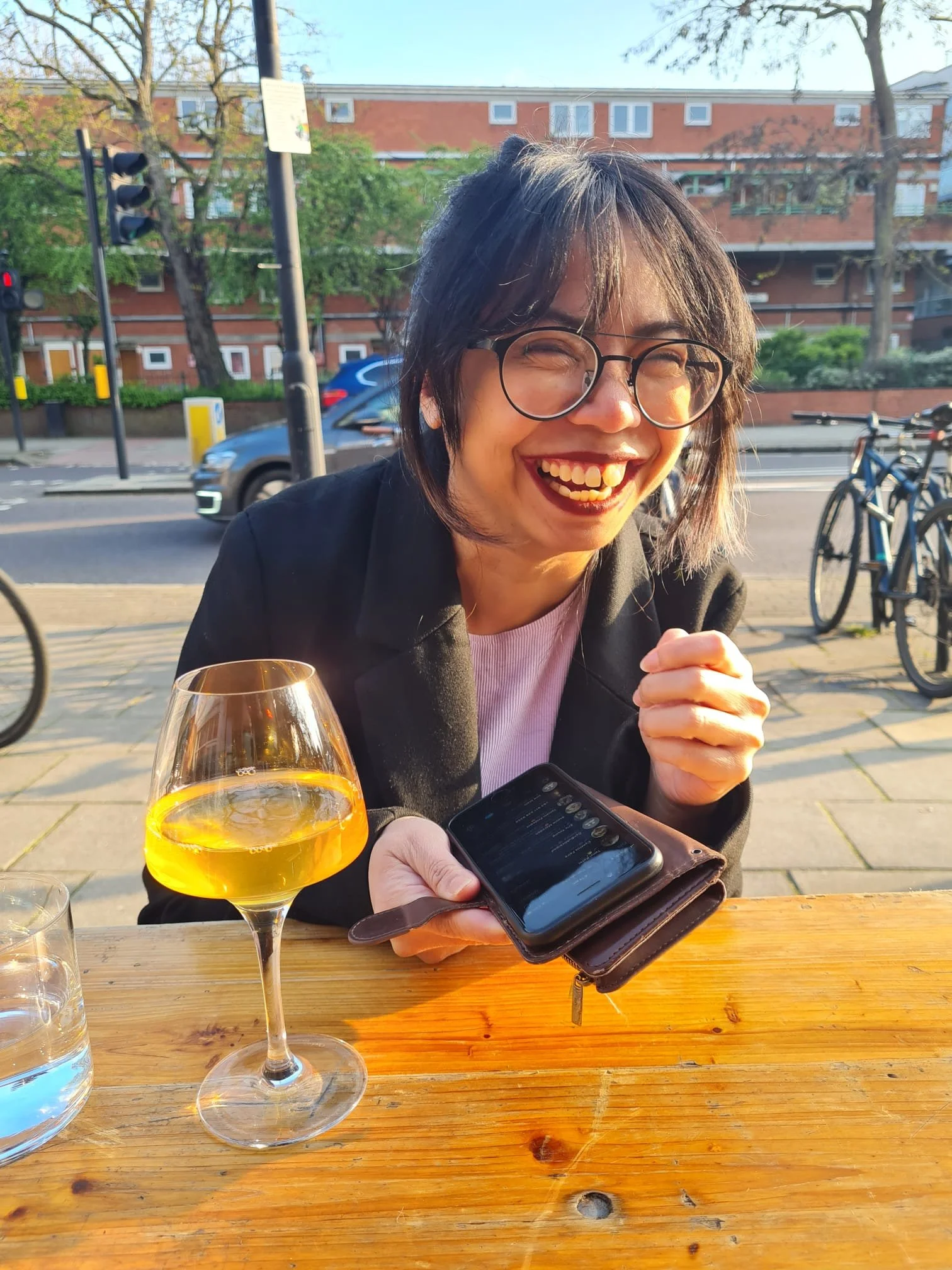 A woman with glasses smiling outdoors at a table, holding a smartphone, with a glass of yellow drink, a glass of water, and bicycles in the background.