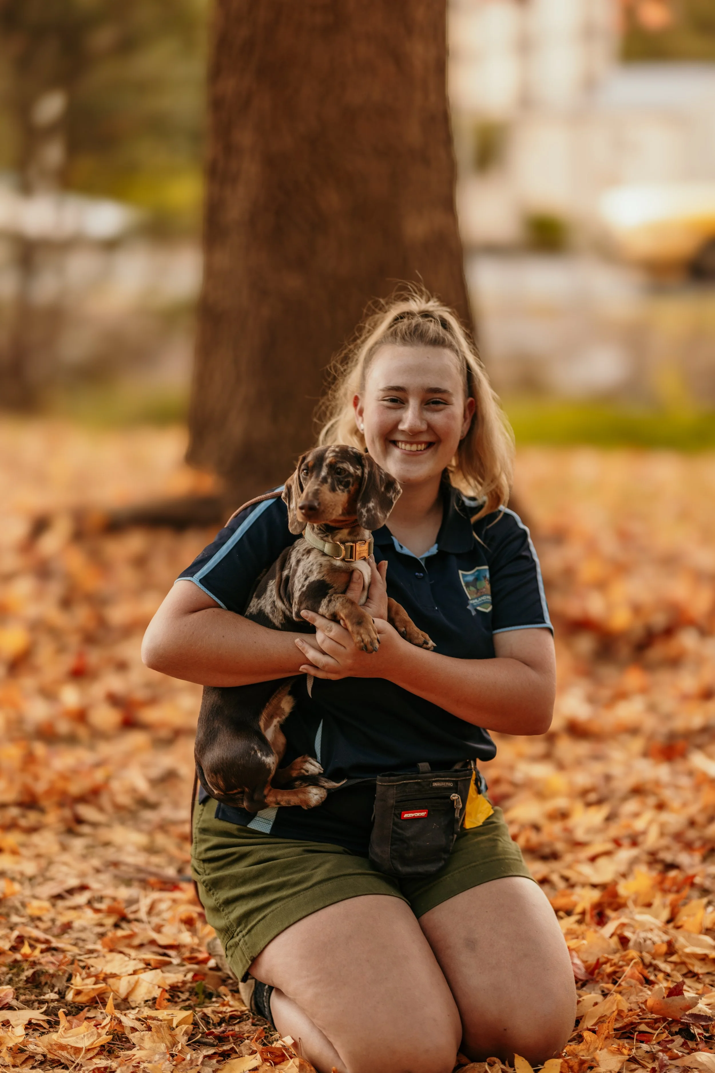 A young woman kneeling on fallen autumn leaves in a park, holding a dachshund puppy. She is smiling and wearing a navy blue shirt with a patch on the chest, olive green shorts, and a waist pouch, in front of a large tree with more leaves on the ground.