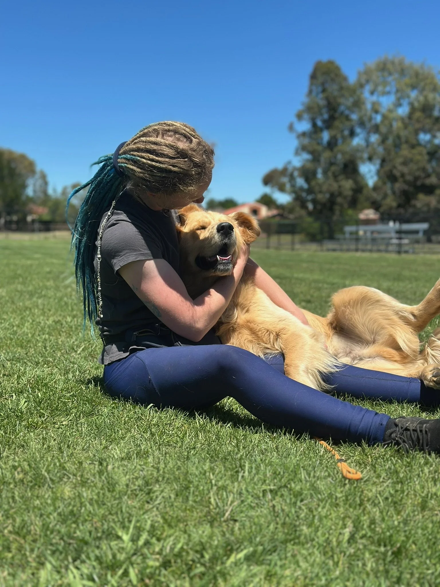 Person with long, dreadlocked hair and dark clothing hugging a golden retriever dog while sitting on green grass at a park on a sunny day.