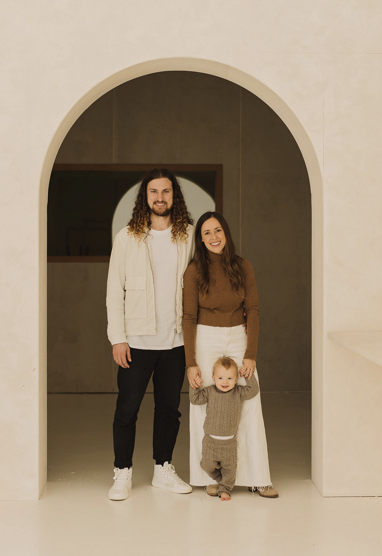 A family of four standing in an arched doorway. The father has long curly hair and is wearing black pants and a white jacket. The mother has long brown hair and is wearing white pants and a brown sweater. Their young child is standing in front, holding the mother's hand, wearing a beige sweater and pants, smiling at the camera.