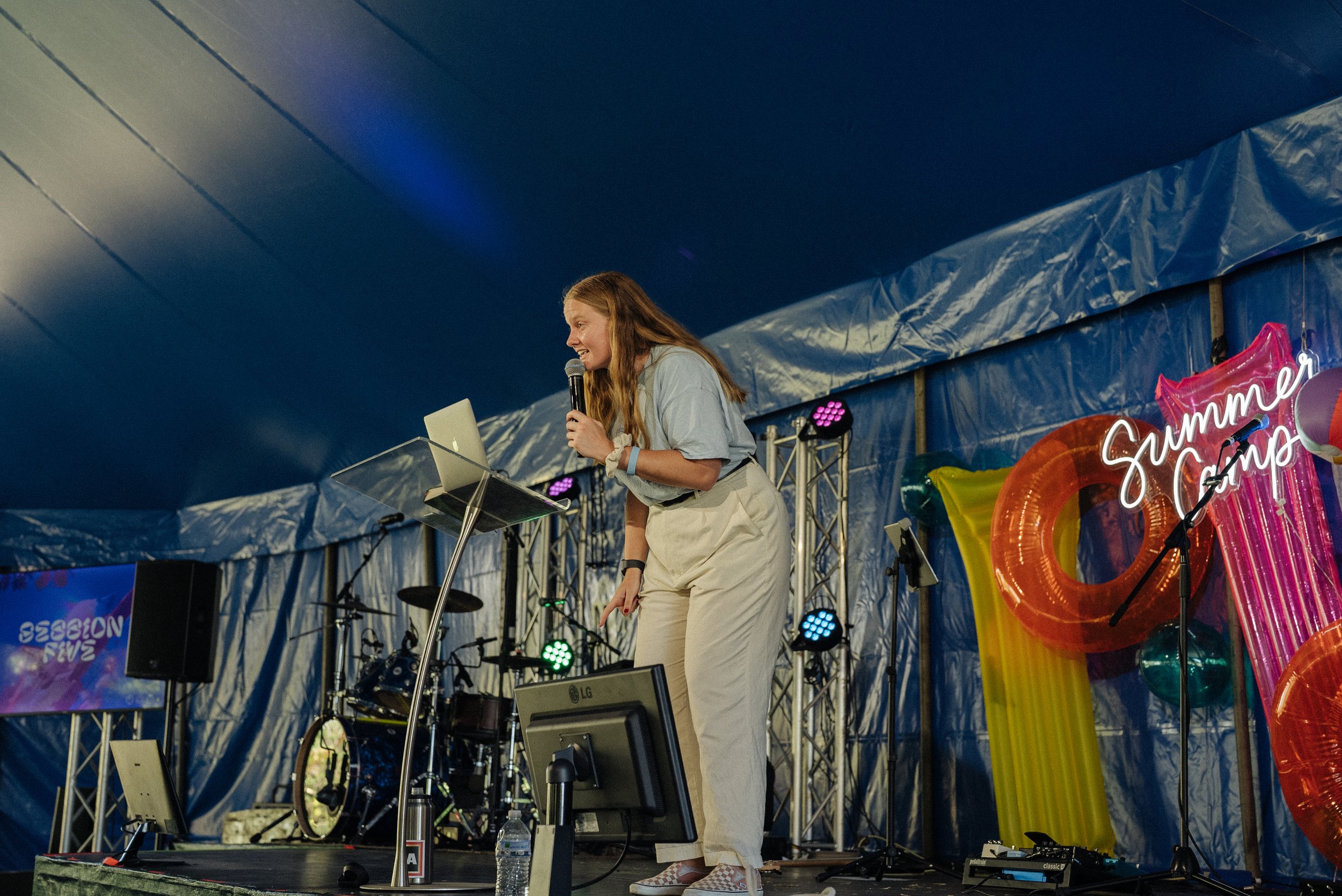 A young woman with long red hair stands on stage, speaking into a microphone, with a table holding a laptop and bottles of water in front of her. She is dressed in a light t-shirt and beige pants. The backdrop includes colorful balloons and a neon sign that says 'Summer Camps' under a blue tent ceiling.