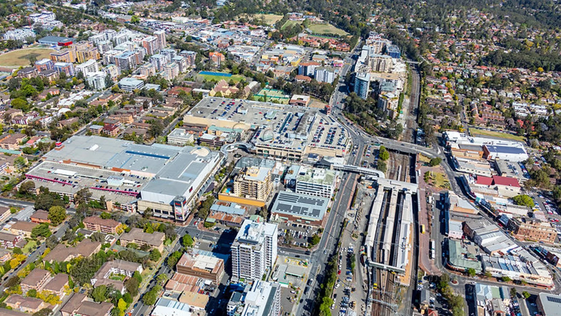 Aerial view of a cityscape showing commercial and residential buildings, parking lots, green sports fields, and roads with vehicles.